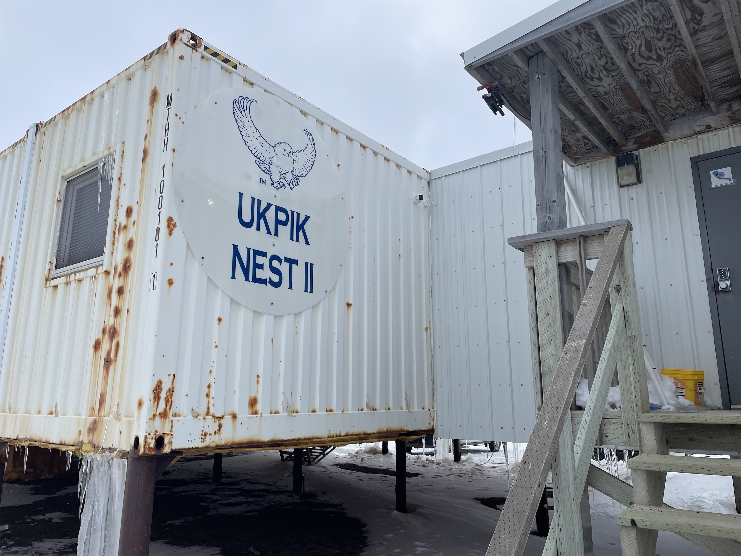 A sign featuring the words "Ukpik Nest II" hangs on the side of a white, rusting metal building elevated on posts.