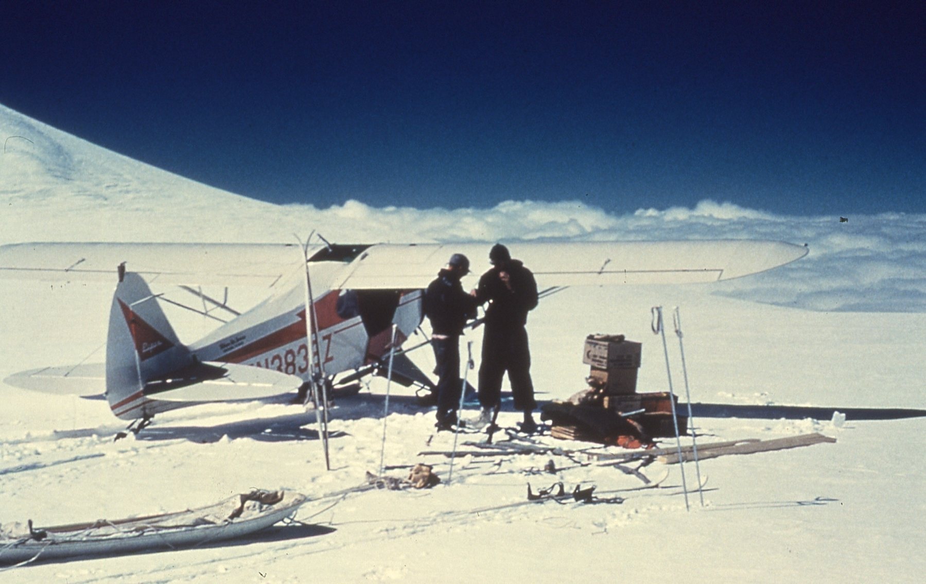 Two men stand next to a small aircraft on a snowy plane.