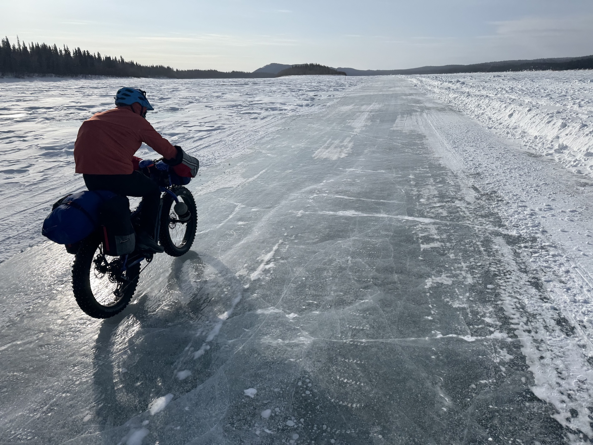 A person in a blue helmet and an orange windbreaker rides a fat-tired bike on a ice road plowed on a river, with drifted snow to the left and a snow berm to the right. Tall evergreens line the riverbank to the left, and hills rise in the distance.