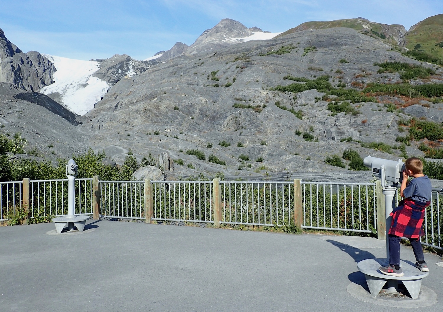 A boy looks at a small glacier in a mountain using a telescope mounted on a concrete viewing platform.