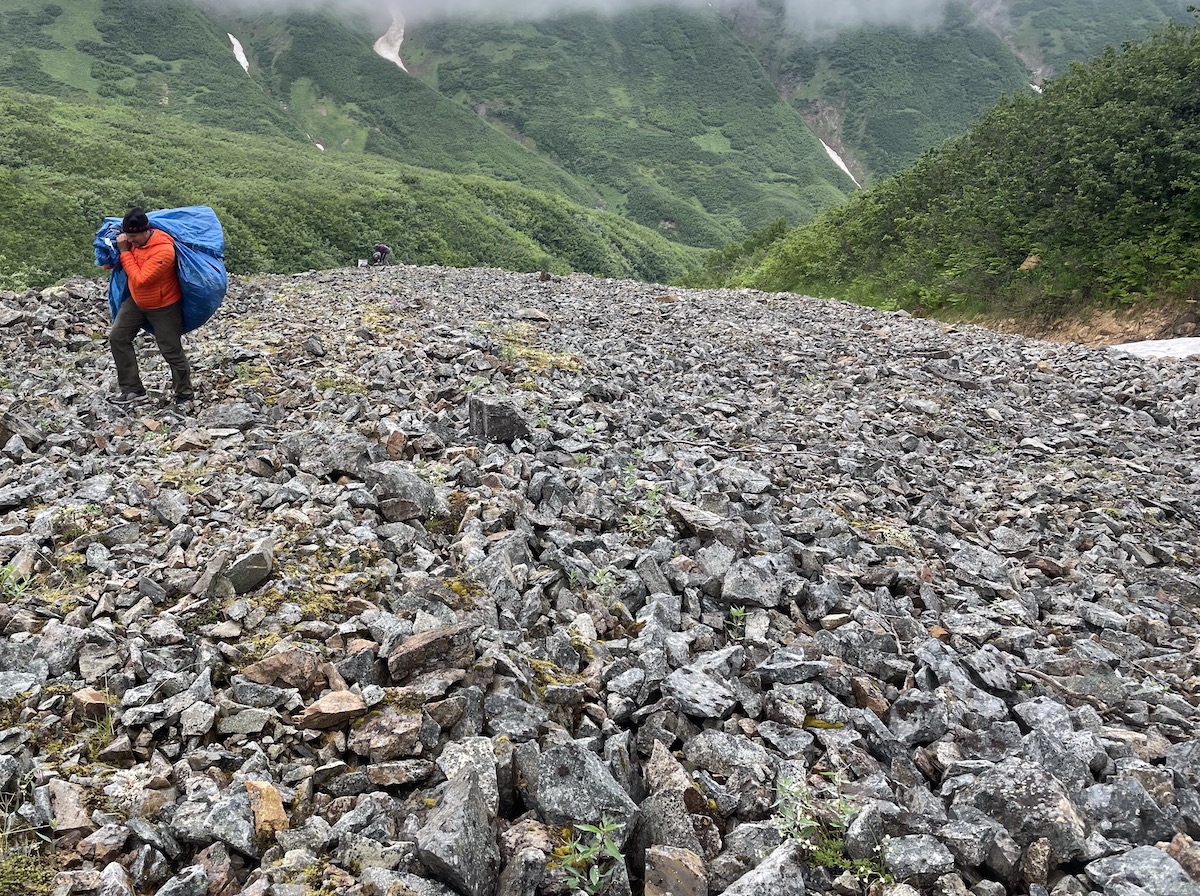 A man hauls a bulging blue tarp across a jumbled slope of rocks. In the background are steep, lush green hillsides with snow drifts lingering in gullies.