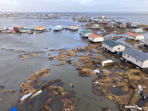 Aerial view of the coastal Alaska village of Kipnuk severely flooded, with homes on stilts surrounded by brown floodwater, debris-strewn land, and displaced vegetation stretching to the horizon in October 2025.