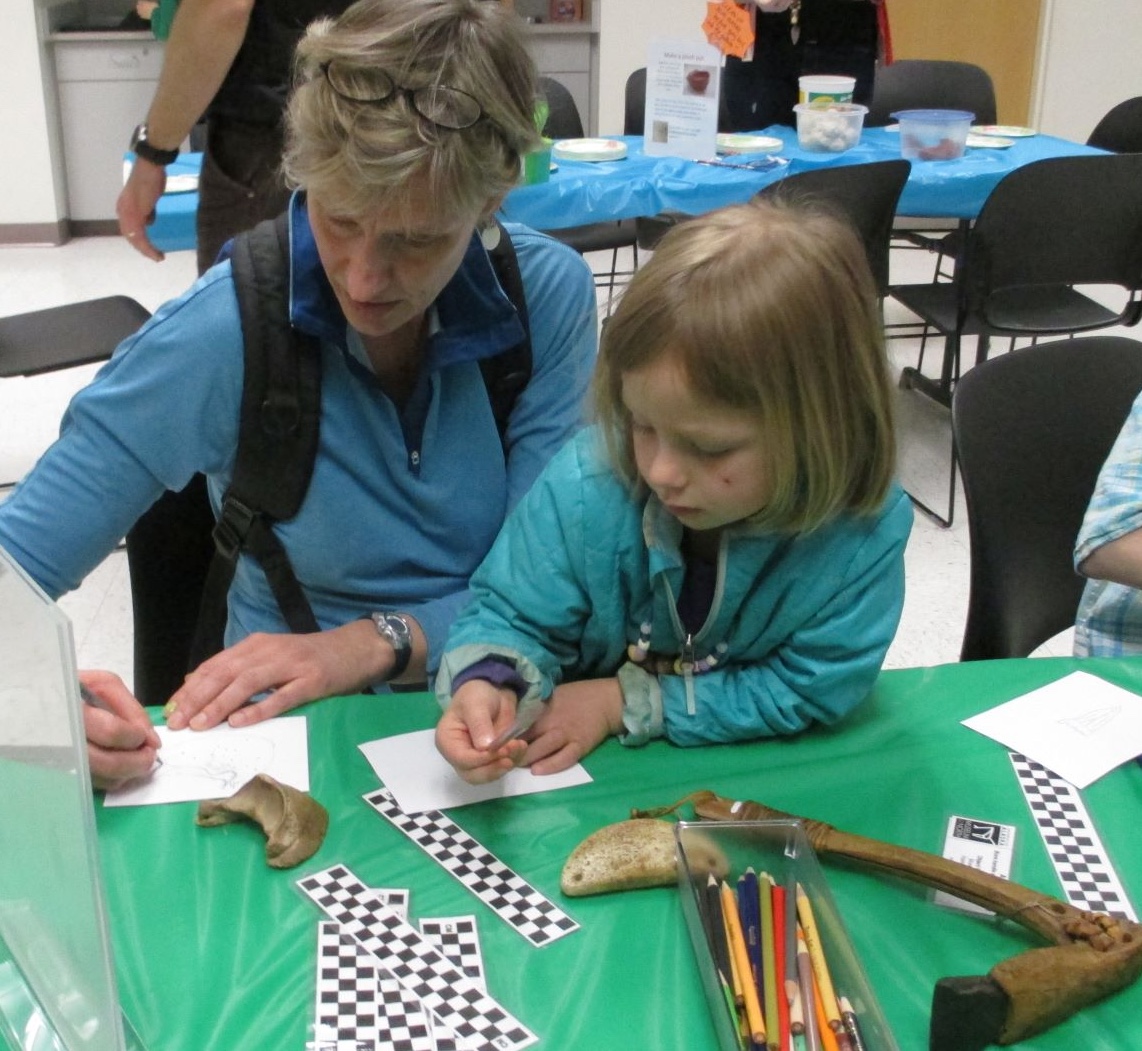 An adult and child sit at a table with measuring grids, paper and pencils. They are looking at and sketching a bone artifact.