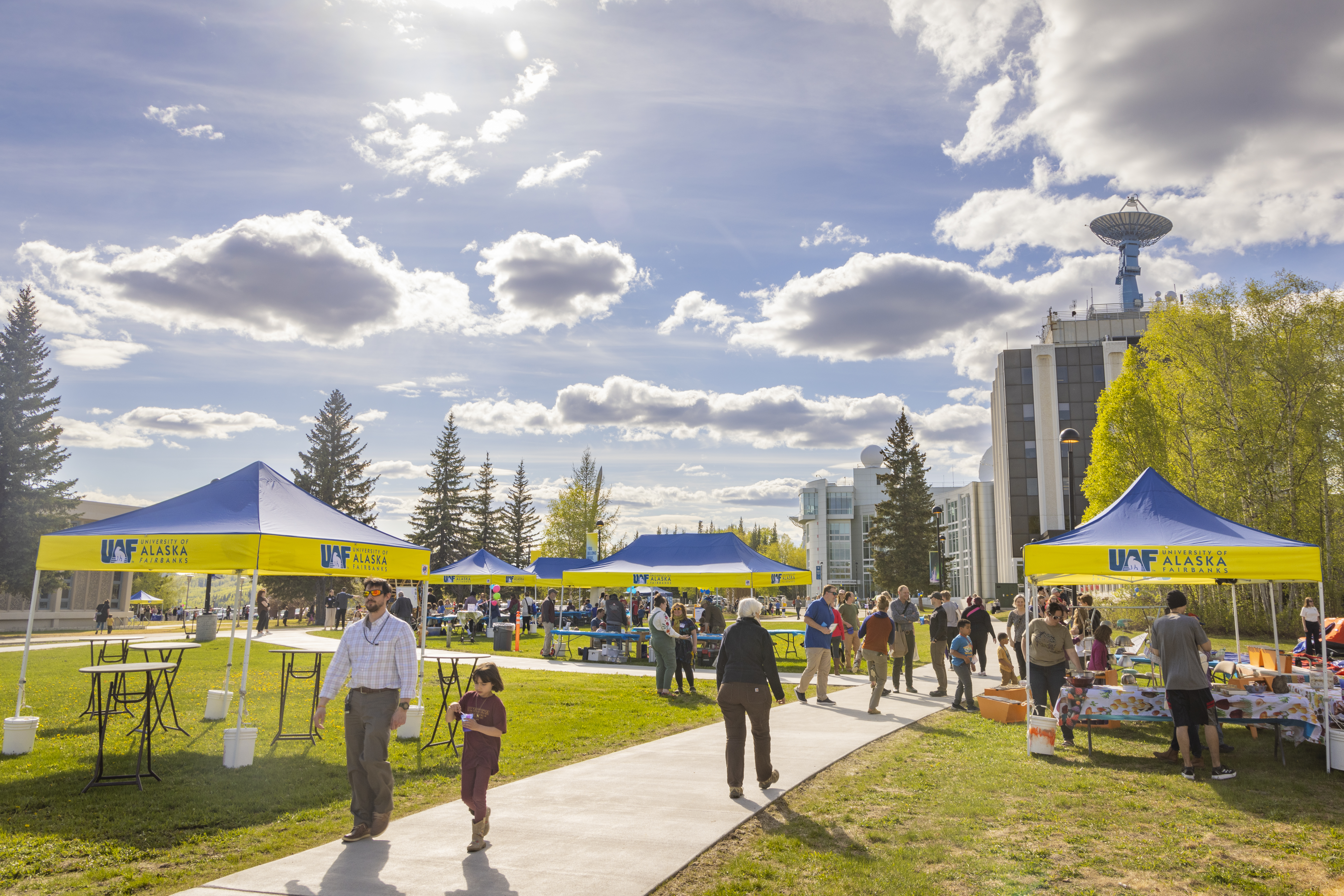 Visitors walk among UAF-branded blue and gold tents set up along a campus pathway during the Arctic Research Open House on the University of Alaska Fairbanks campus in May 2025, with the Elvey Building and a satellite dish visible in the background under a sunny sky.