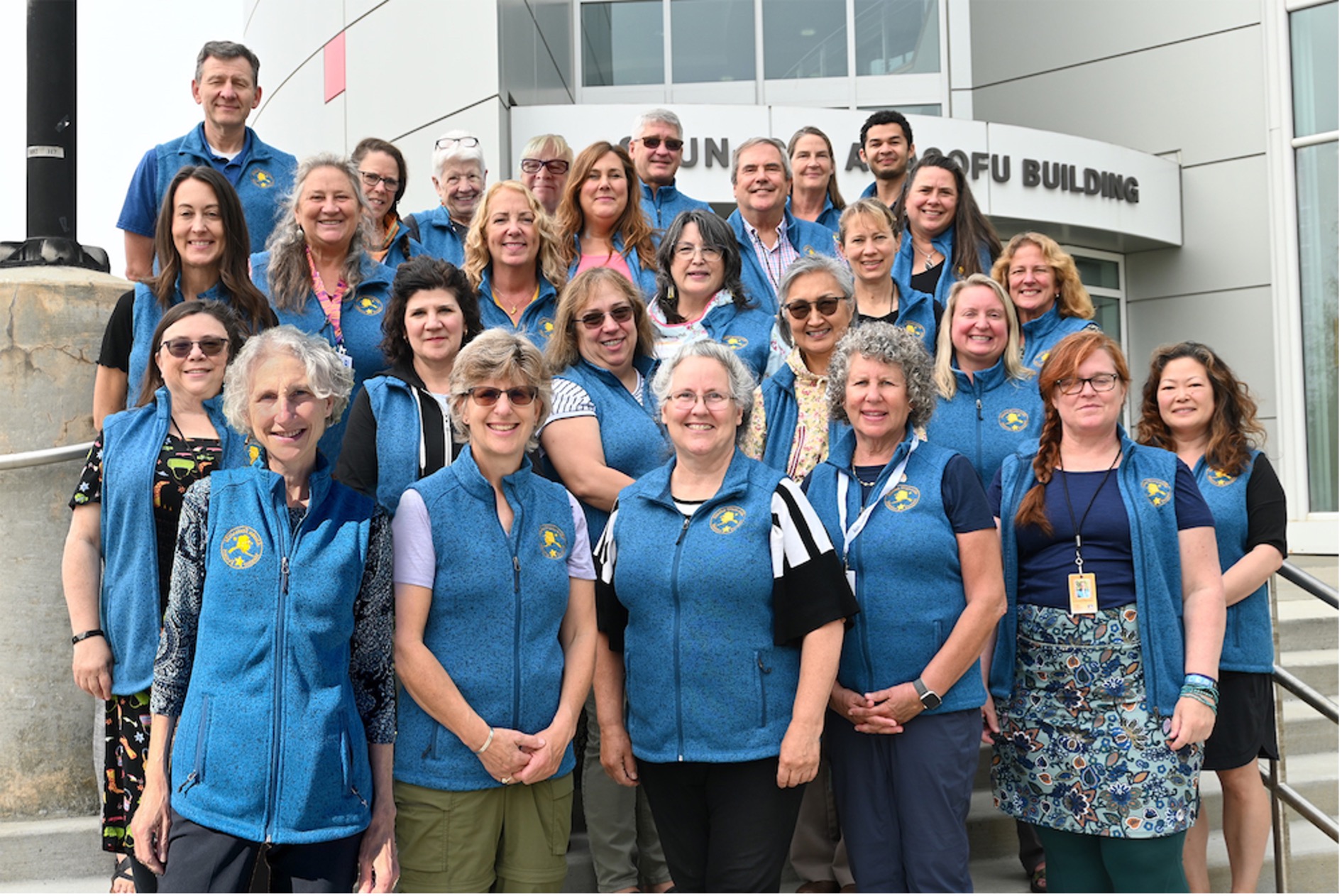 A group of 27 adults wearing matching light blue vests with gold patches gathered for a group photo on the steps of the Akasofu Building at UAF