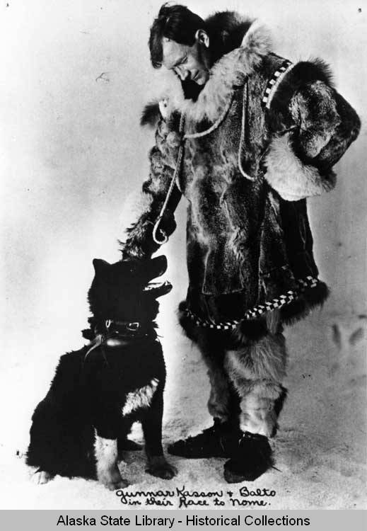 A man in a fur coat pets a dog in a posed black and white photograph.