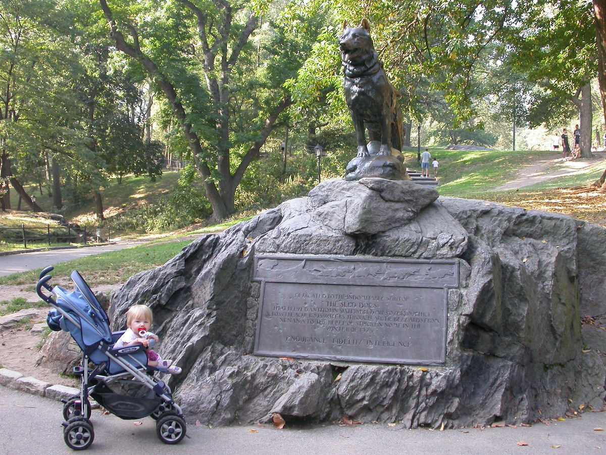 A young girl sits in a stroller in front of an outdoor sculpture of a dog.