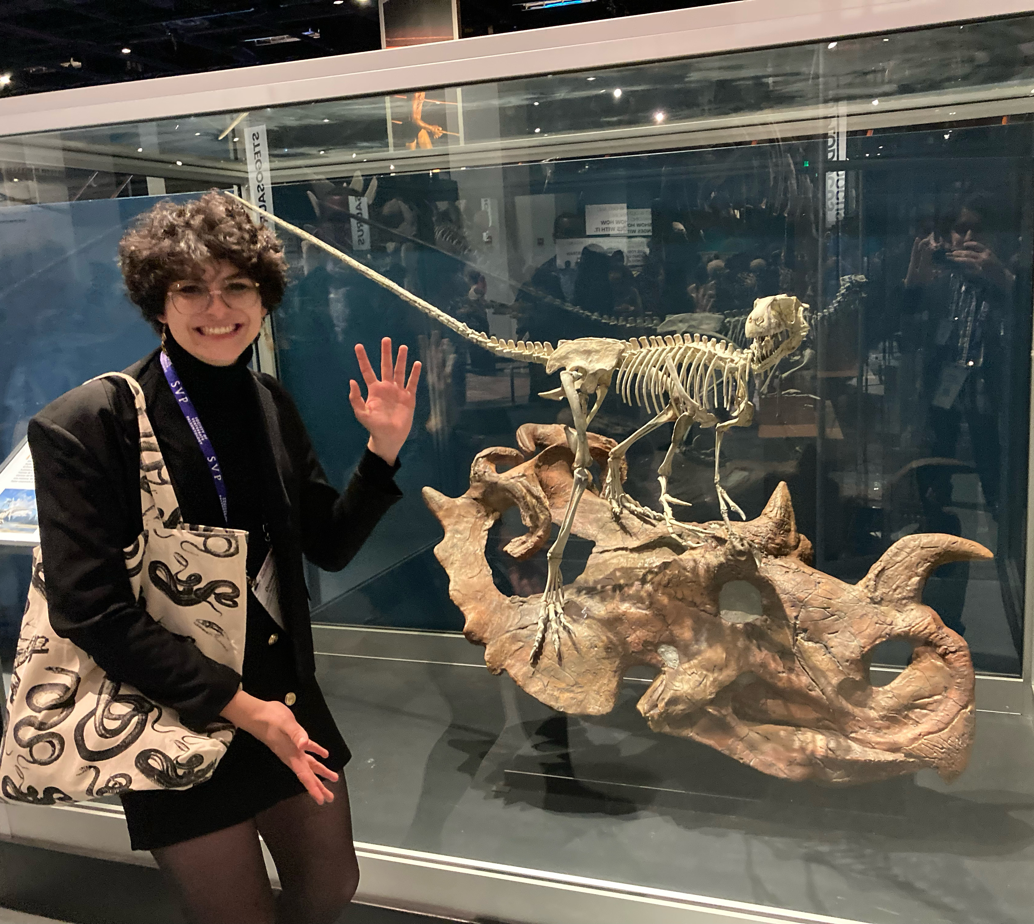 Daniela Barrera-Guevara poses in front of a museum case containing the skeleton of a small dinosaur atop the skull of a larger, horned dinosaur.