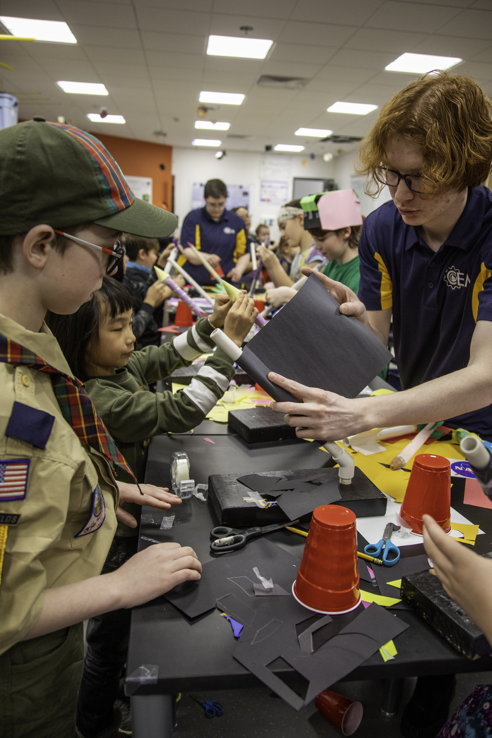 A college student helps a Boy Scout and other children construct a paper rocket.