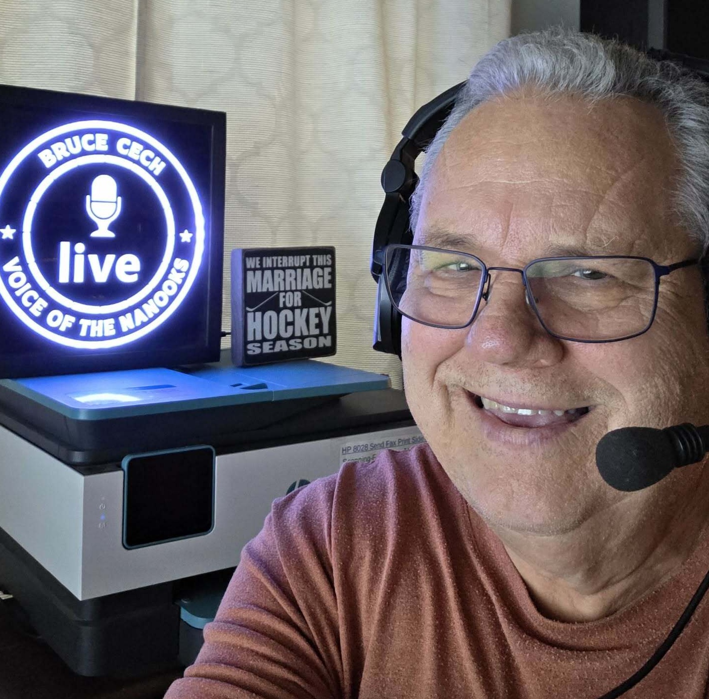 Close-up shot of Bruce Cech in a broadcasting studio wearing a mic, with a lighted sign in the background that reads, "Bruce Cech, Voice of the Nanooks, Live." An unlighted sign says, "We interrupt this marriage for hockey season."