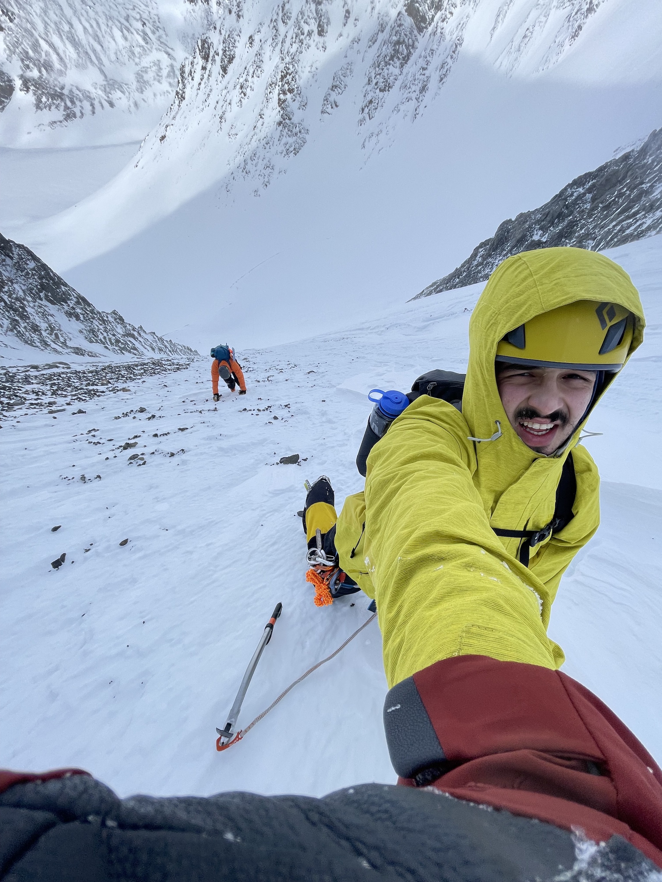 Close-up shot of a mountain climber in a yellow jacket jacket and helmet reaching up towards the camera with another climber lower down the slope
