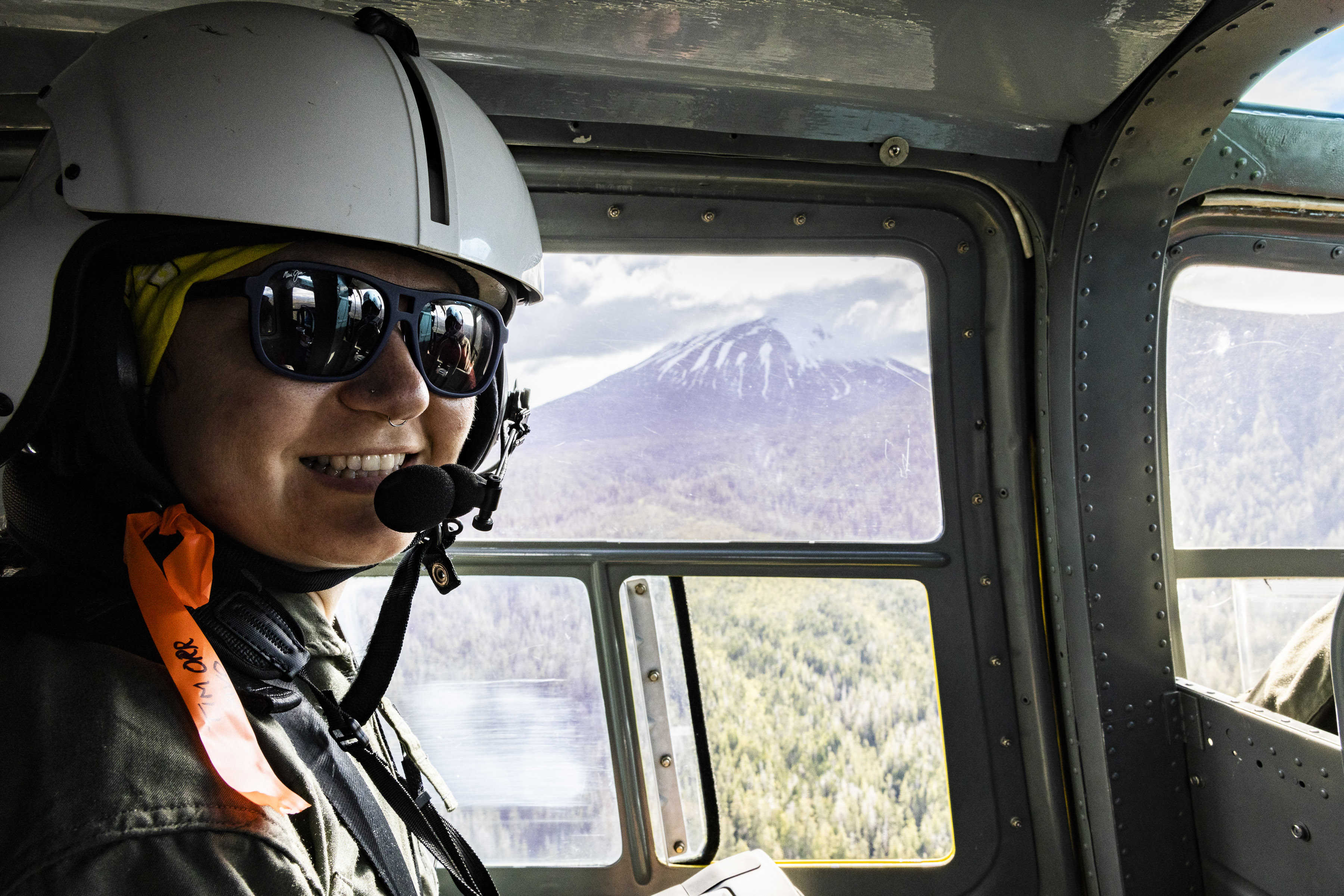 Photo shot from within a small airplane of a woman in a helmet with a headset microphone. Through the window is a mountain with steam rising from the top.