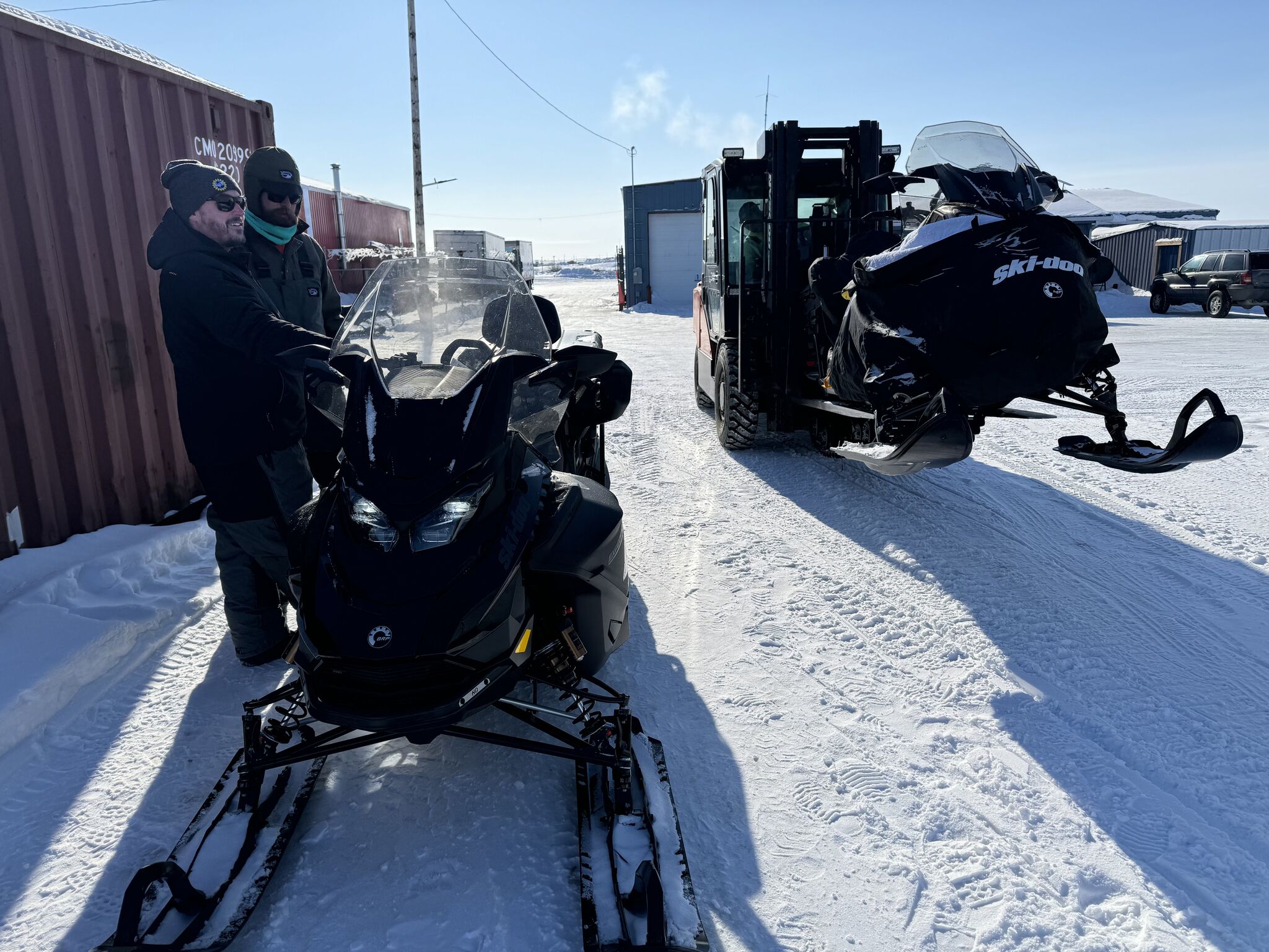 Two men stand beside two snowmachines in a snowy industrial setting.