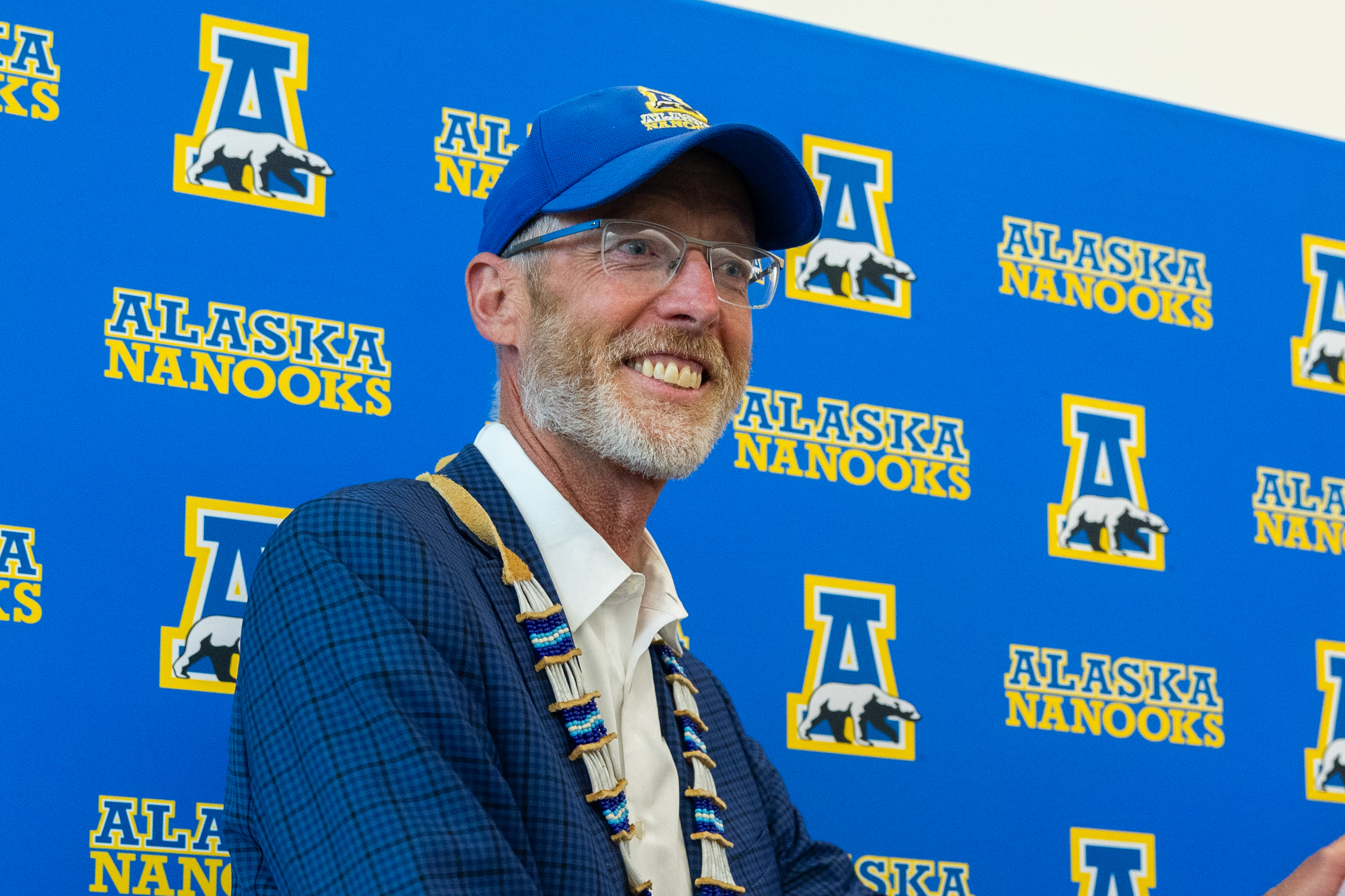 A smiling man wearing a blue suit and blue ball cap stands in front of a blue backdrop with Alaska Nanooks logos