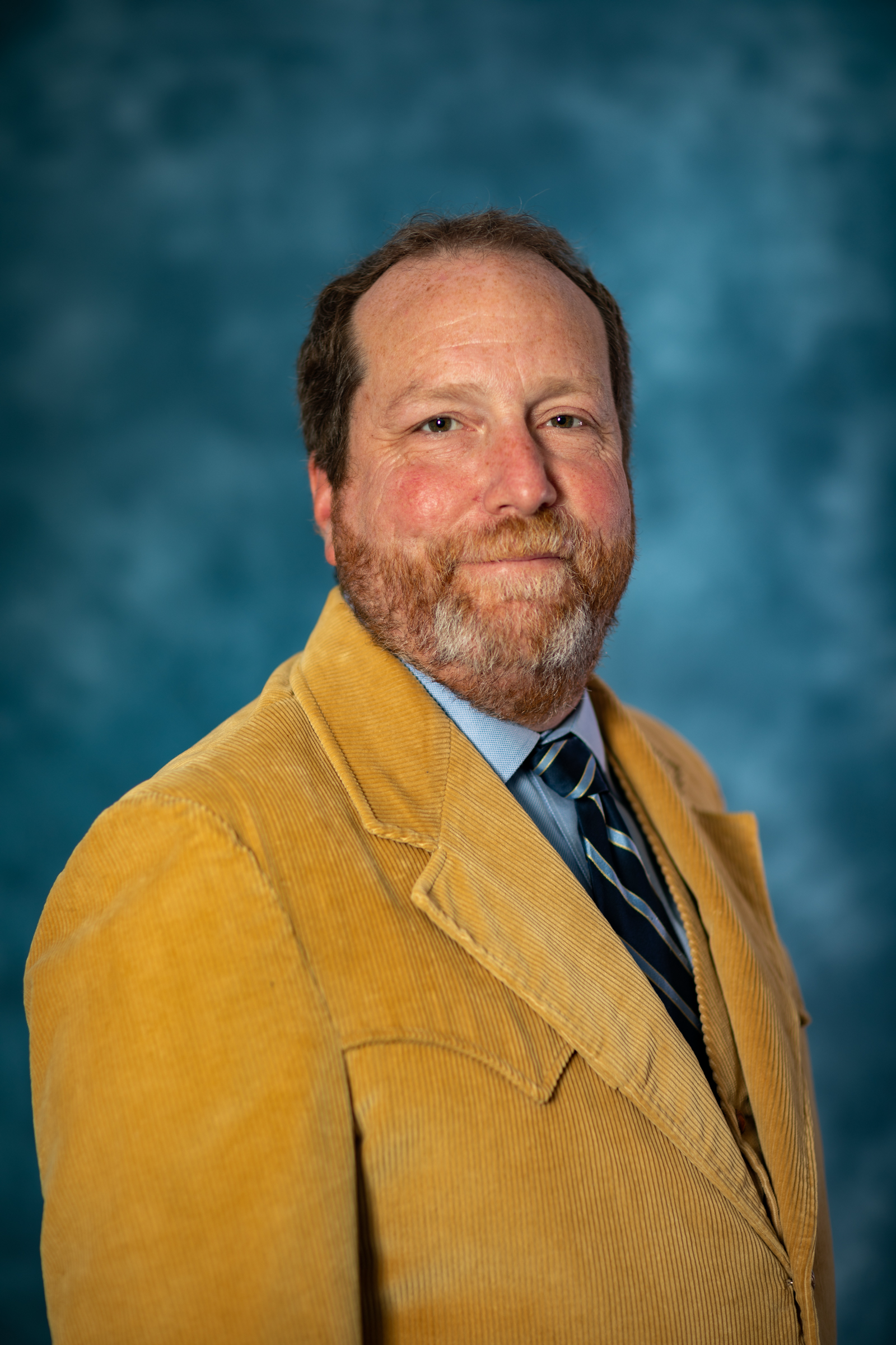 A bearded man wearing a yellow jacket and light blue shirt with a striped tie.