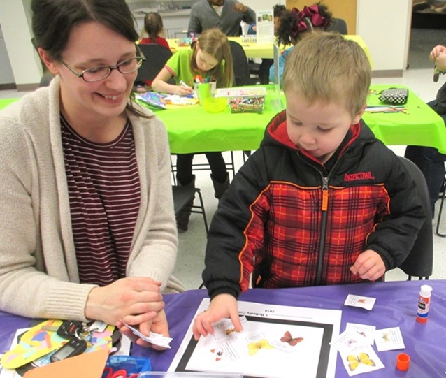 A woman and a young child at an activity table. The child is gluing crayon-colored butterflies to an activity sheet.