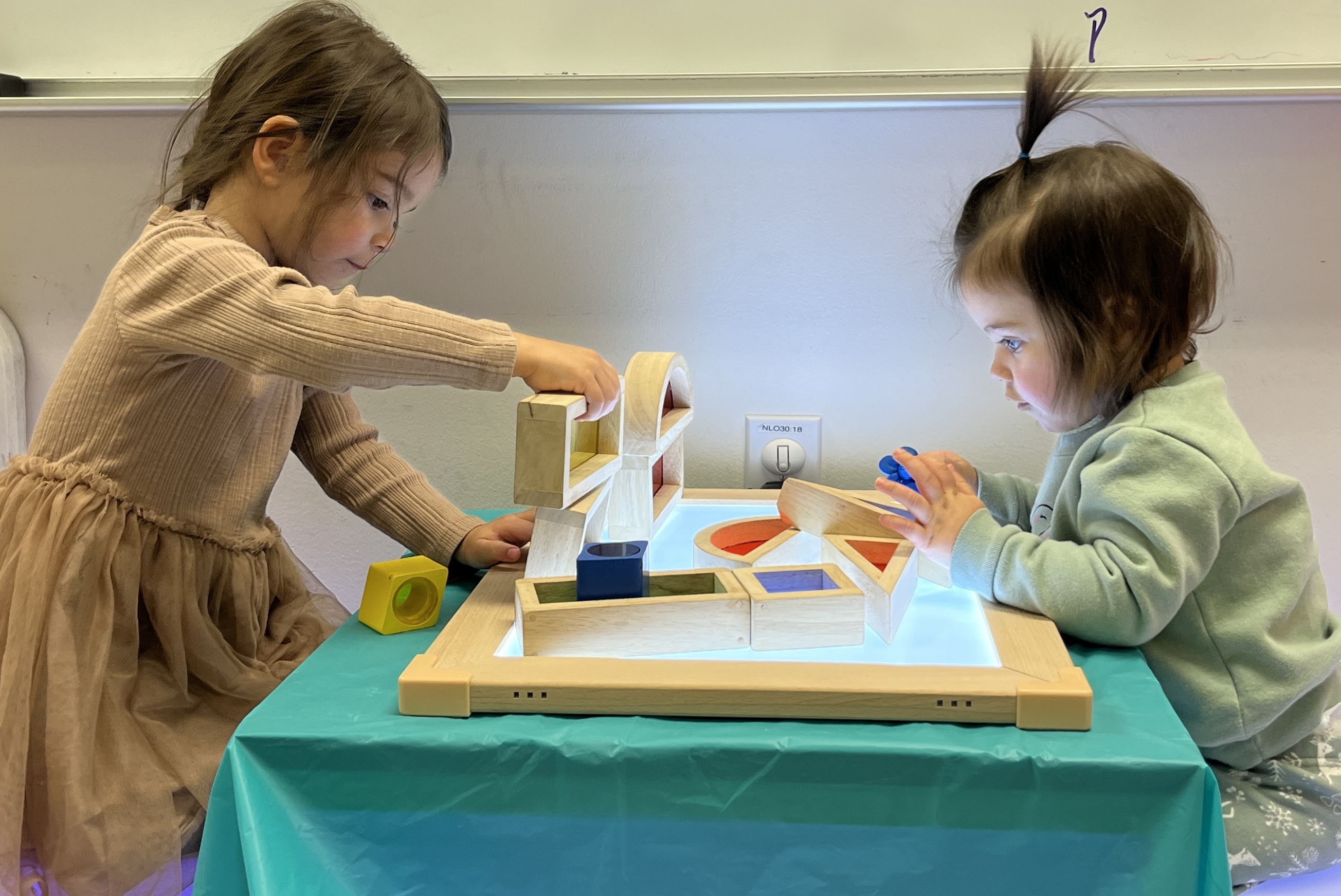 A pair of children sit on opposite sides of a lighted table, playing with hollow wooden geometric shapes.