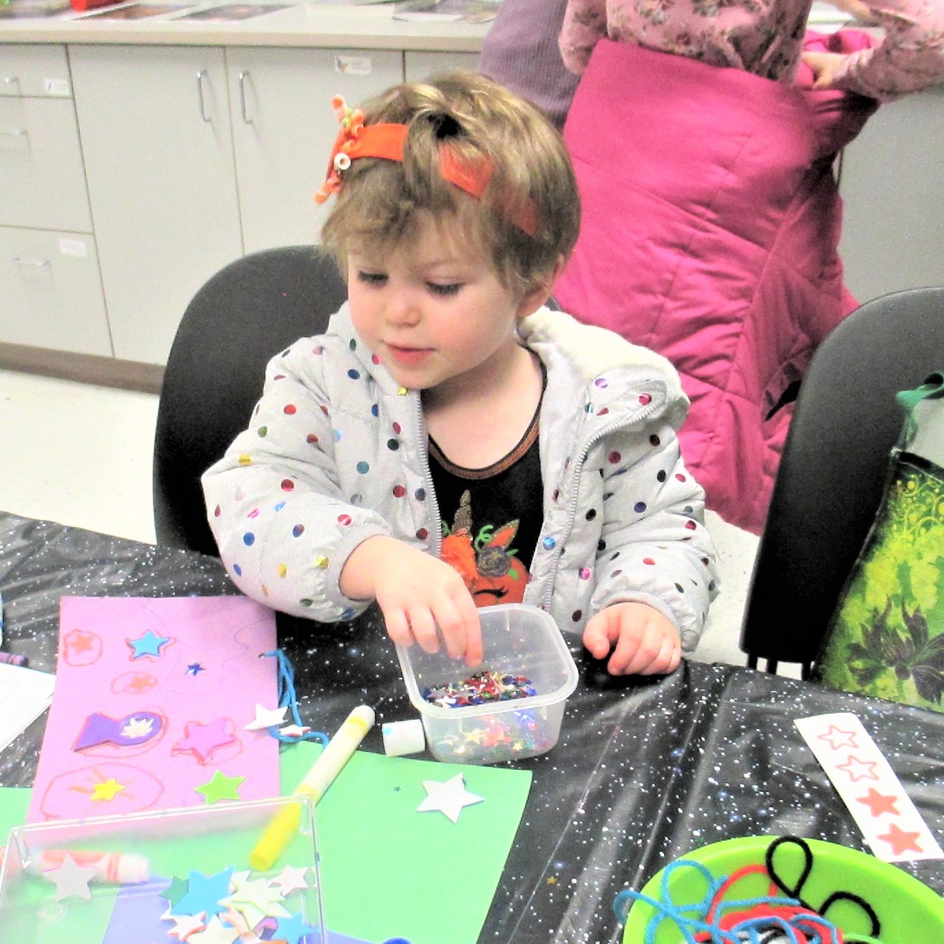 Sitting at a table covered with star-themed tablecloth, a child uses construction paper, crayons, string, glue and stick-on stars to create artwork.