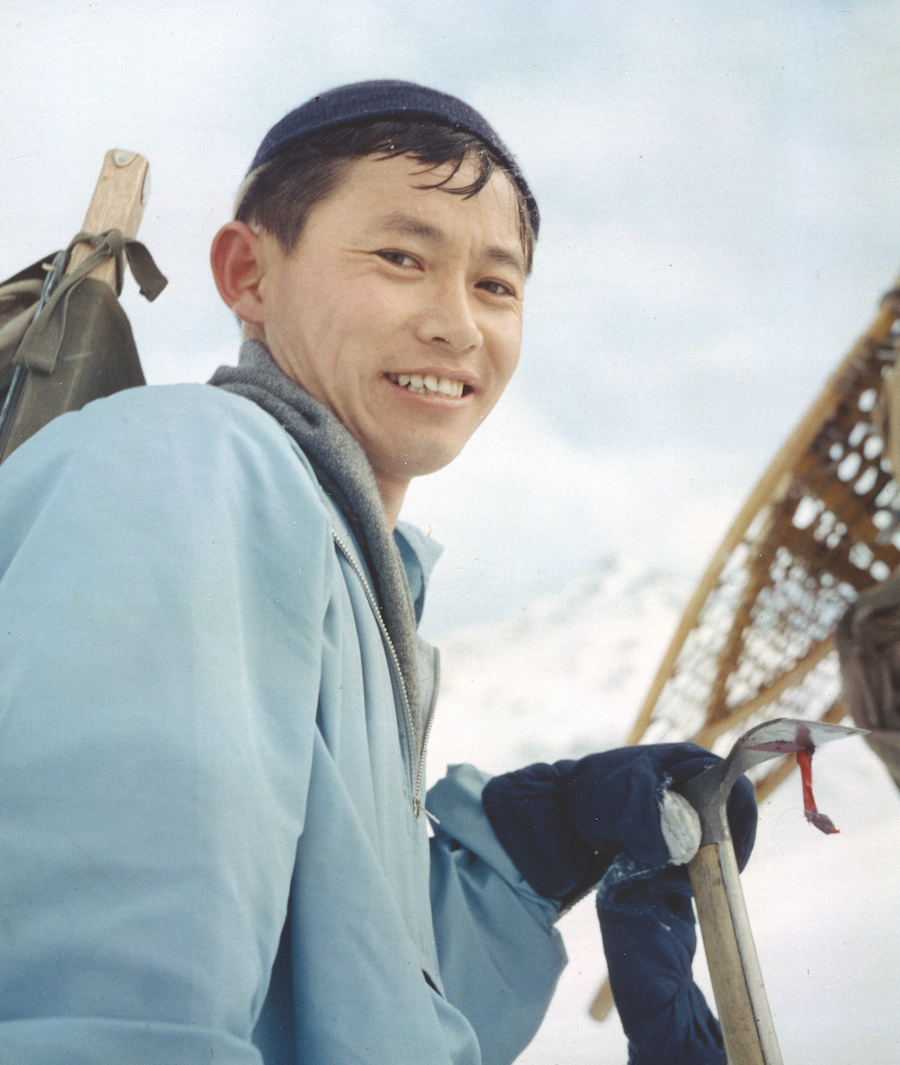A man with a backpack and an ice ax smiles at the camera. A pair of snowshoes and a mountain peak are visible in the background.