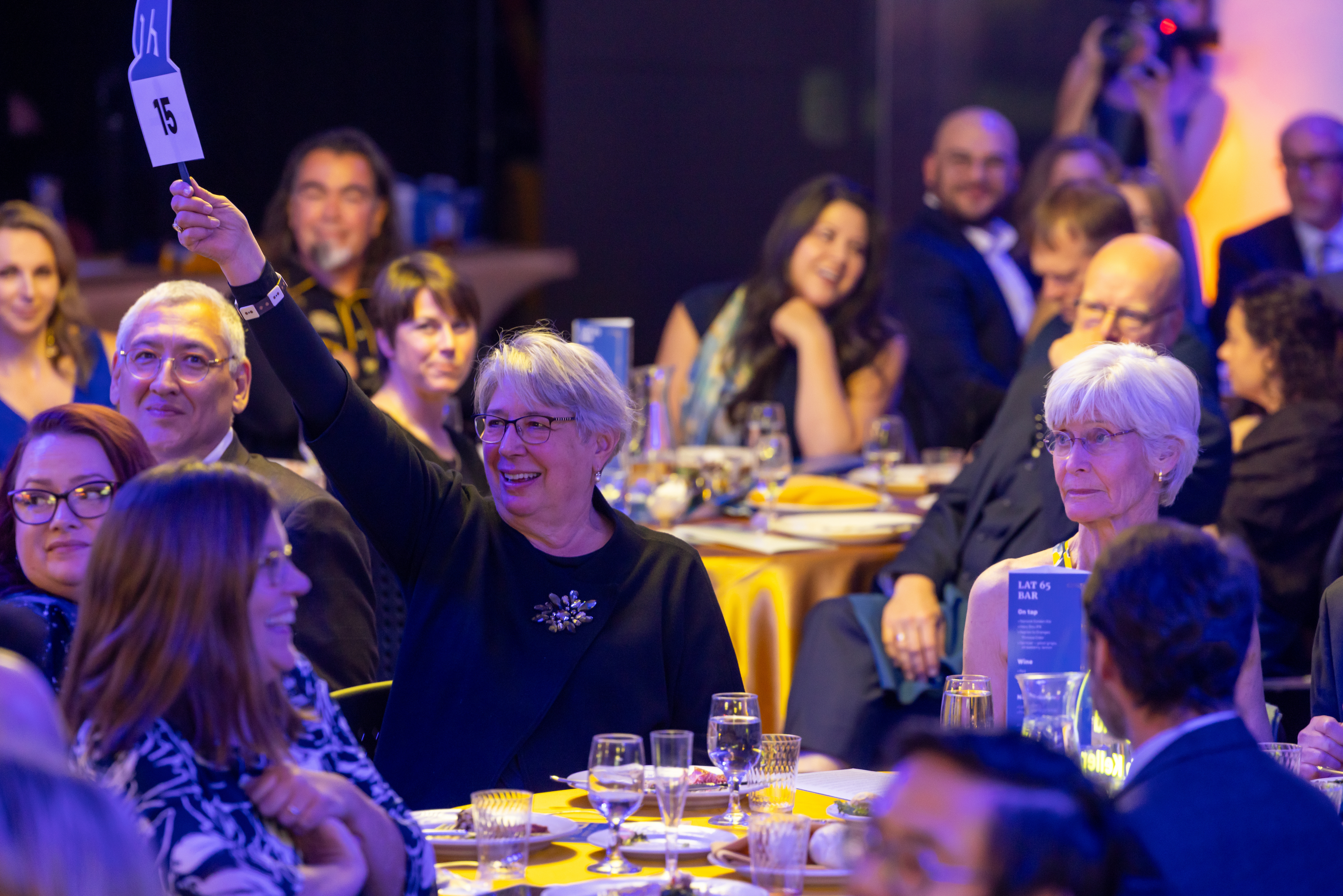A smiling person seated at a banquet table raises a bid paddle. They are surrounded by a large crowd of attendees.