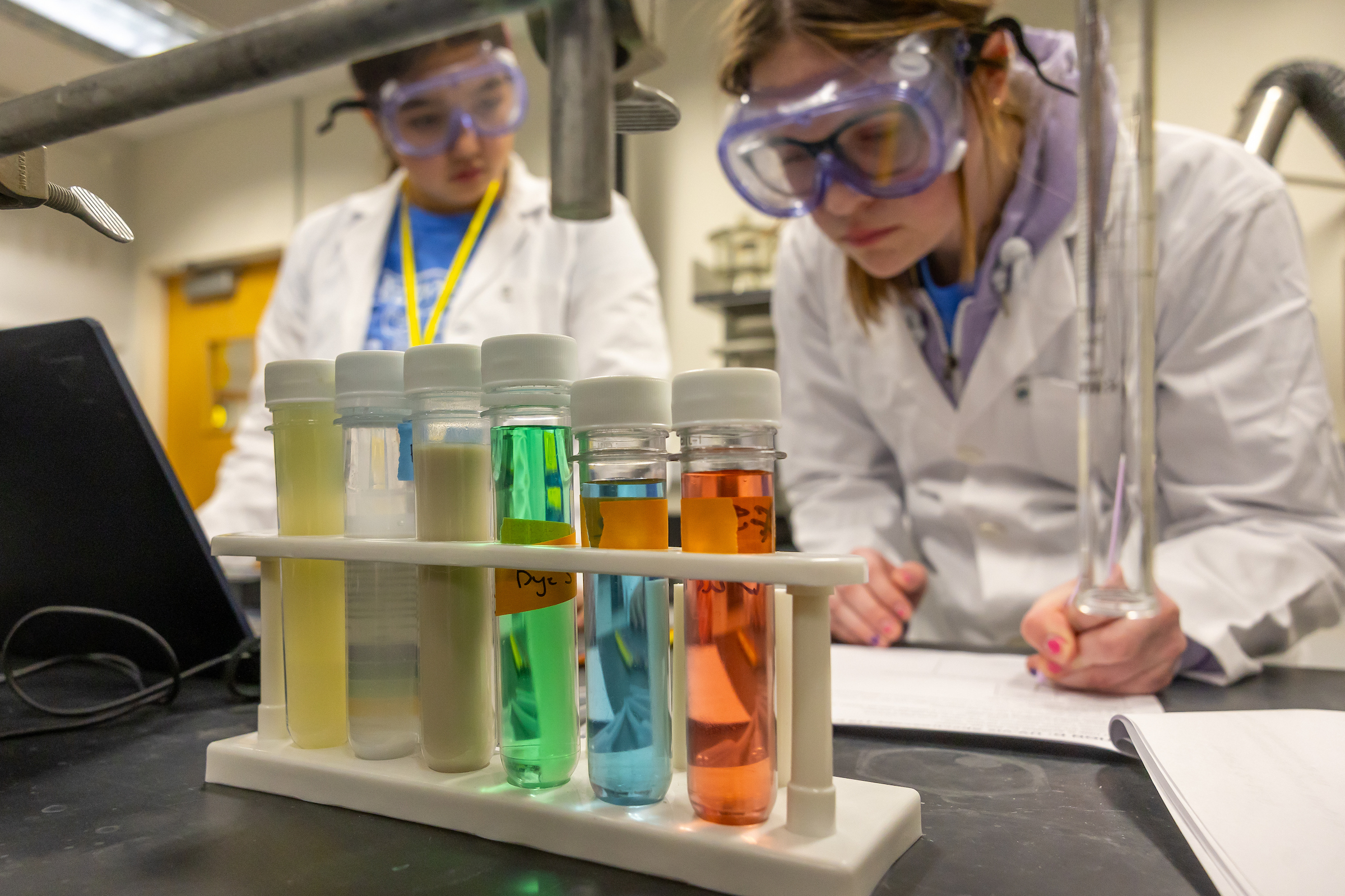 Two middle school students wearing lab coats look at five test tubes filled with liquids of different colors.