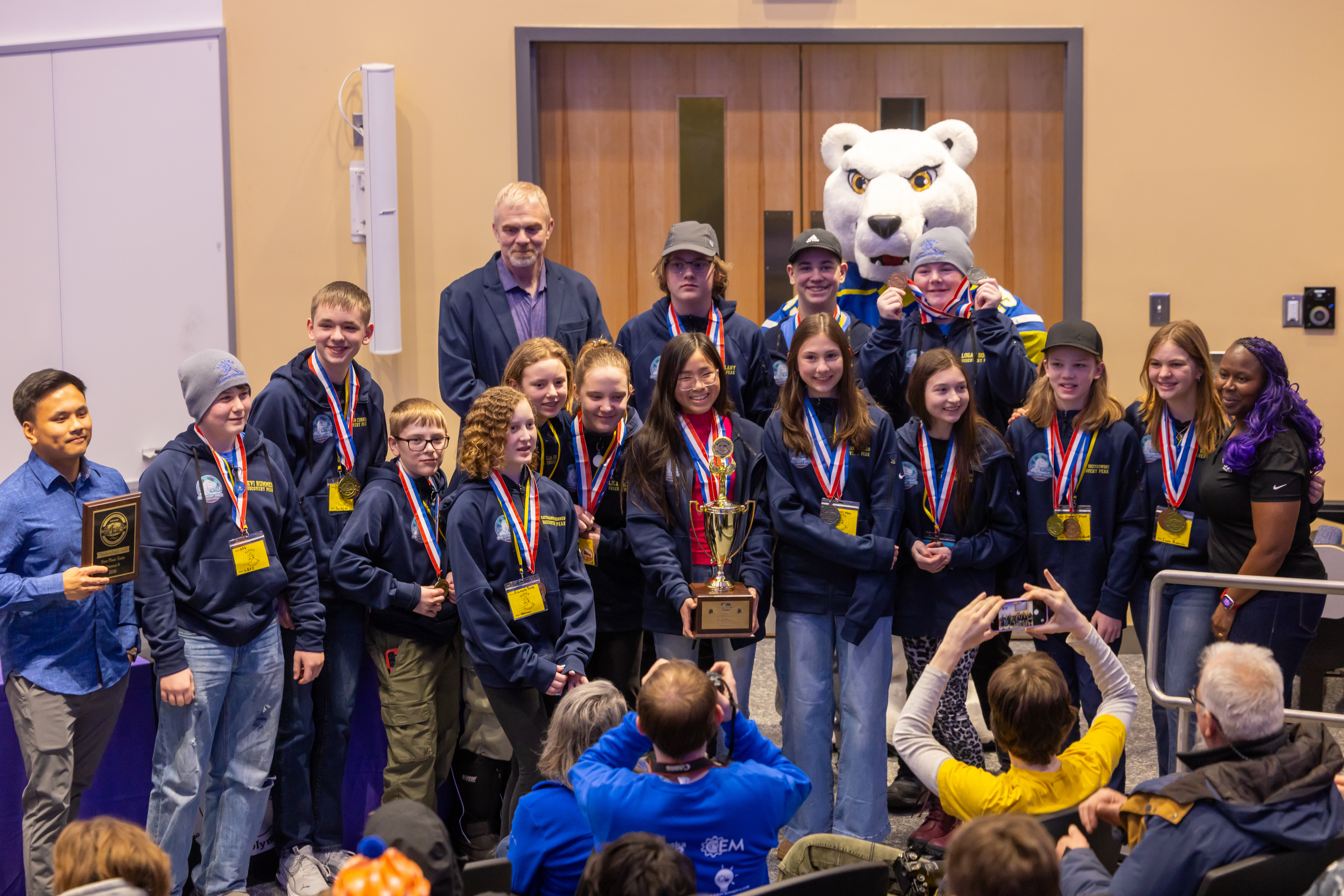 A large group of middle school students wearing first-place medals pose with their trophy and UAF's mascot, Nook, as audience members take photos.