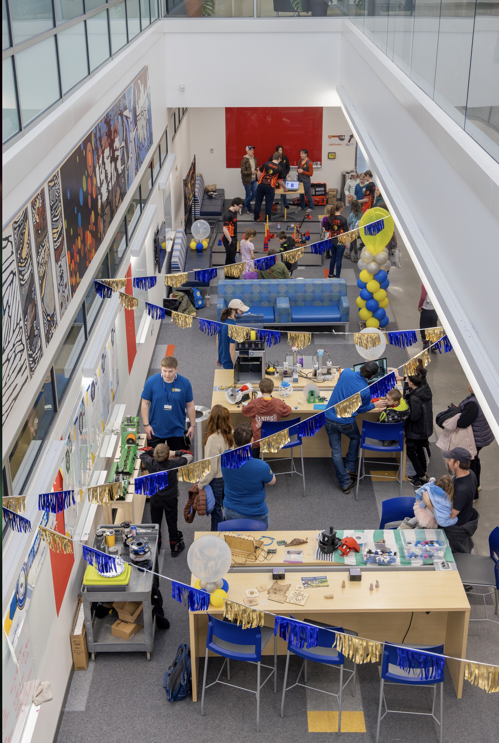 An overhead view shows a bright, two-story atrium filled with long tables set up for hands-on engineering activities. Children and adults gather around demonstrations while UAF students in blue shirts explain projects. Banners and balloons decorate the space, and tools, models and small projects are spread across the tables