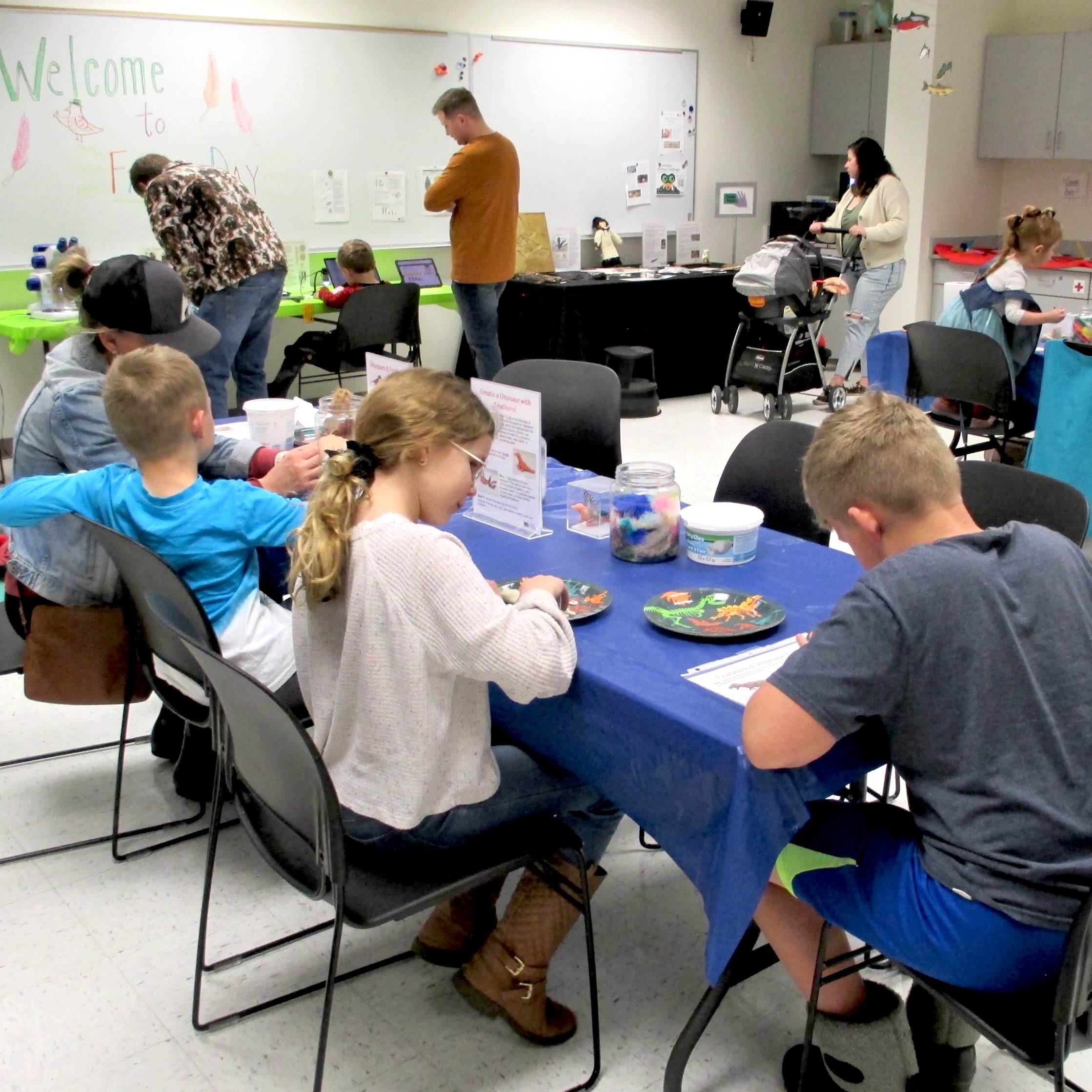 Children and adults work with art materials at tables arranged around a large room. 