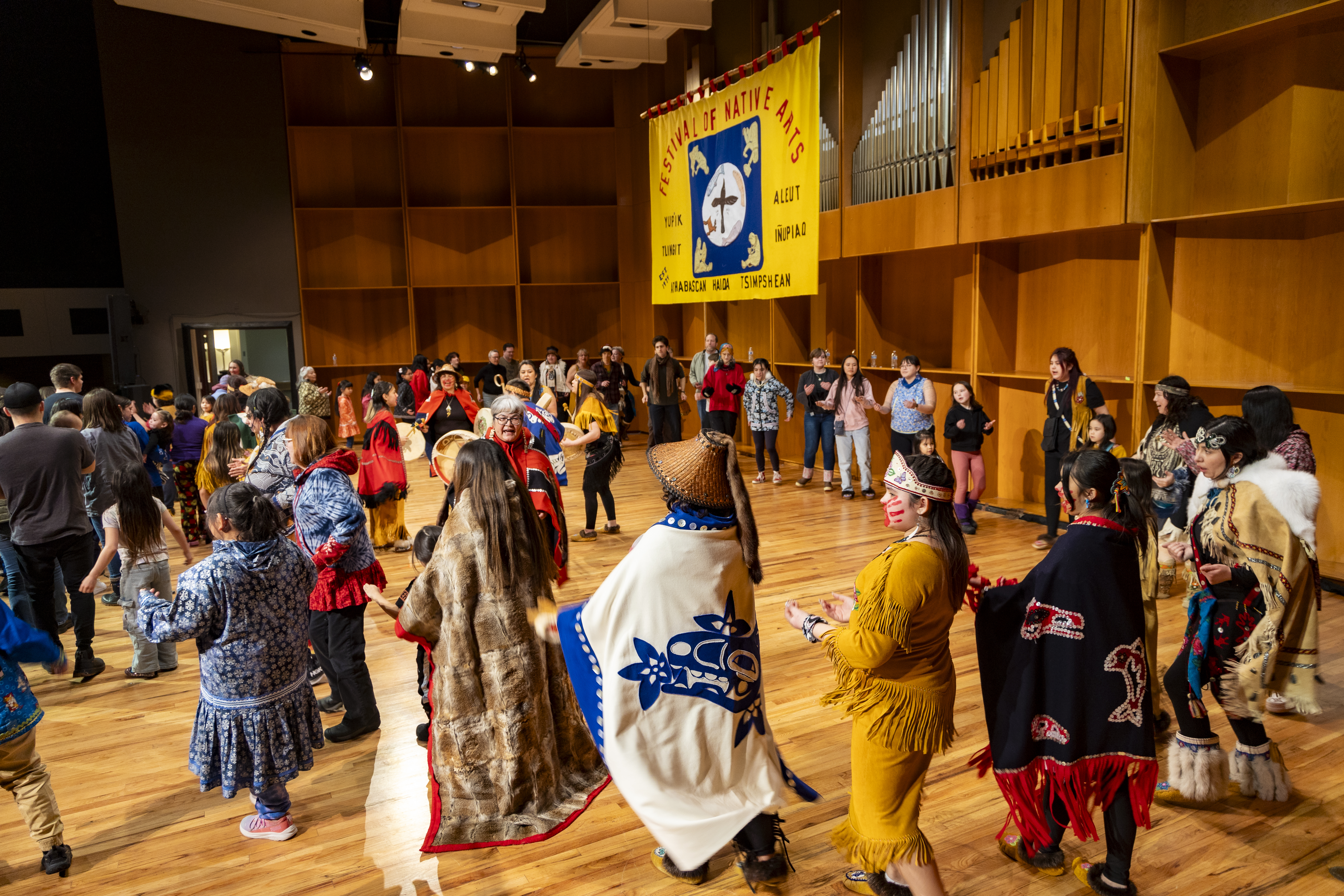 Dozens of people of all ages, many wearing regalia of different tribes, move in a circle around a group of drummers and singers on the stage at the Davis Concert Hall. A large banner hanging from the ceiling includes the words