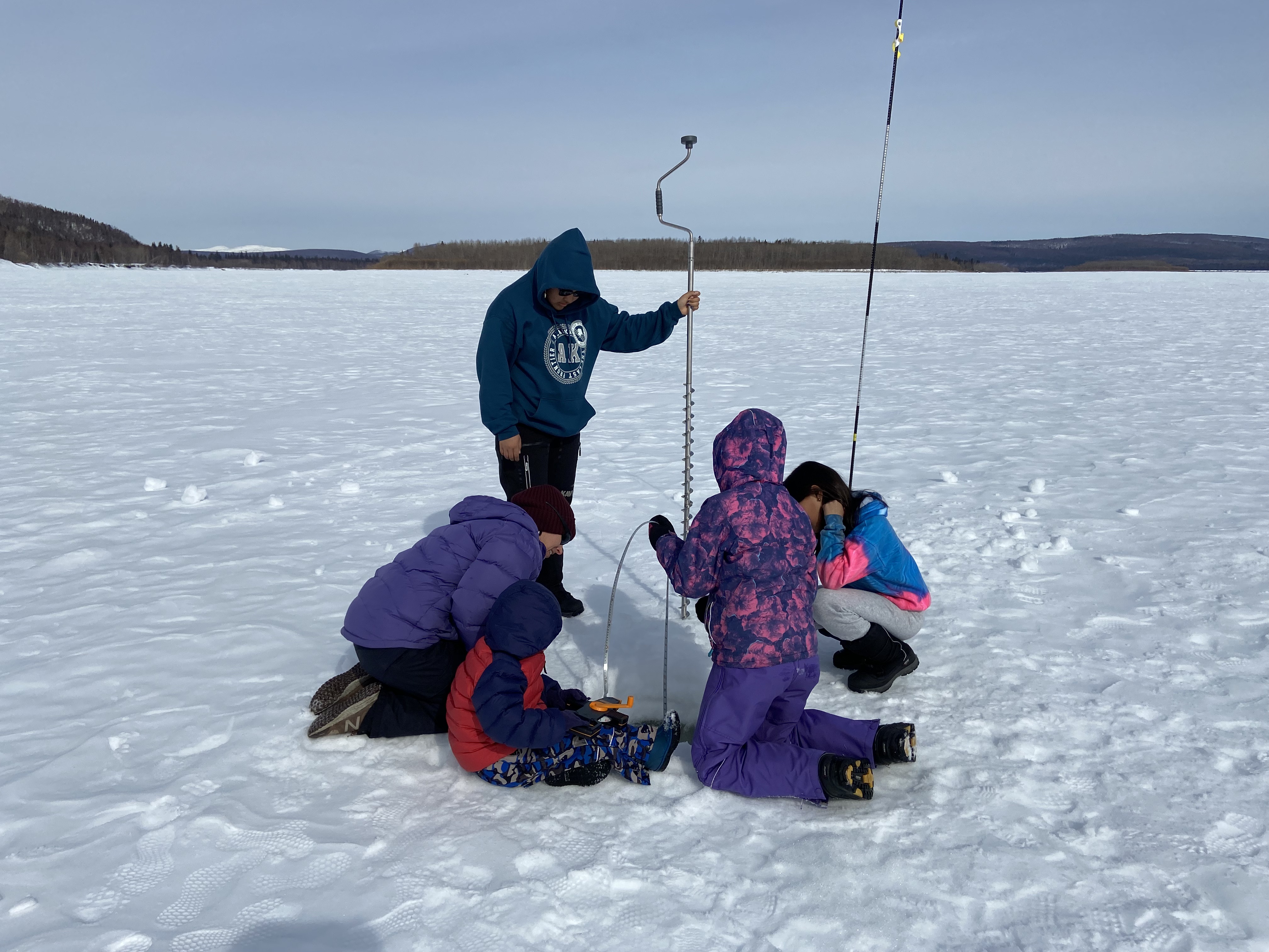 Children and their instructor drill into the ice on a frozen river.