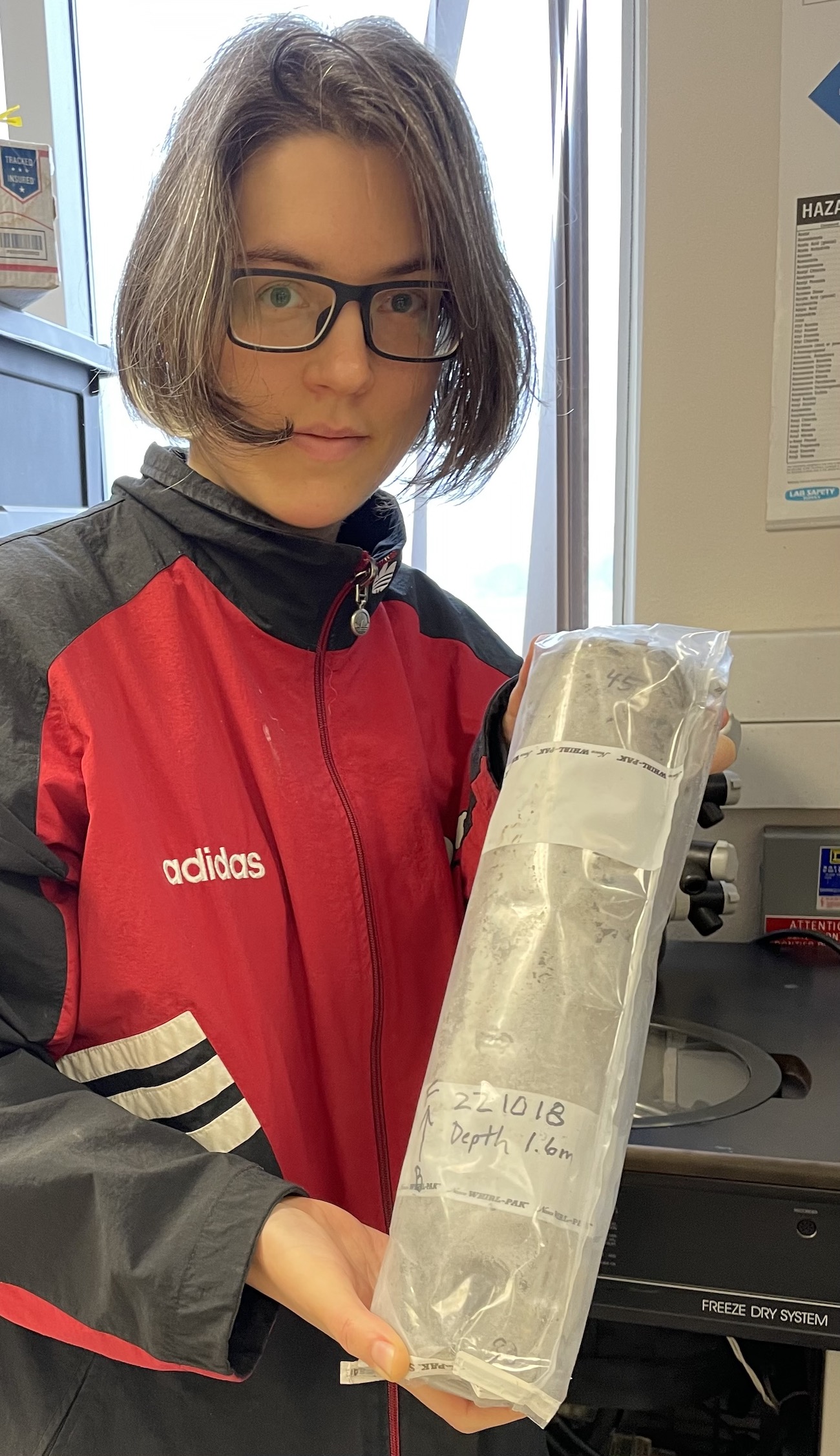 A woman in a red Adidas jacket holds a cylinder of gray soil in a plastic bag.