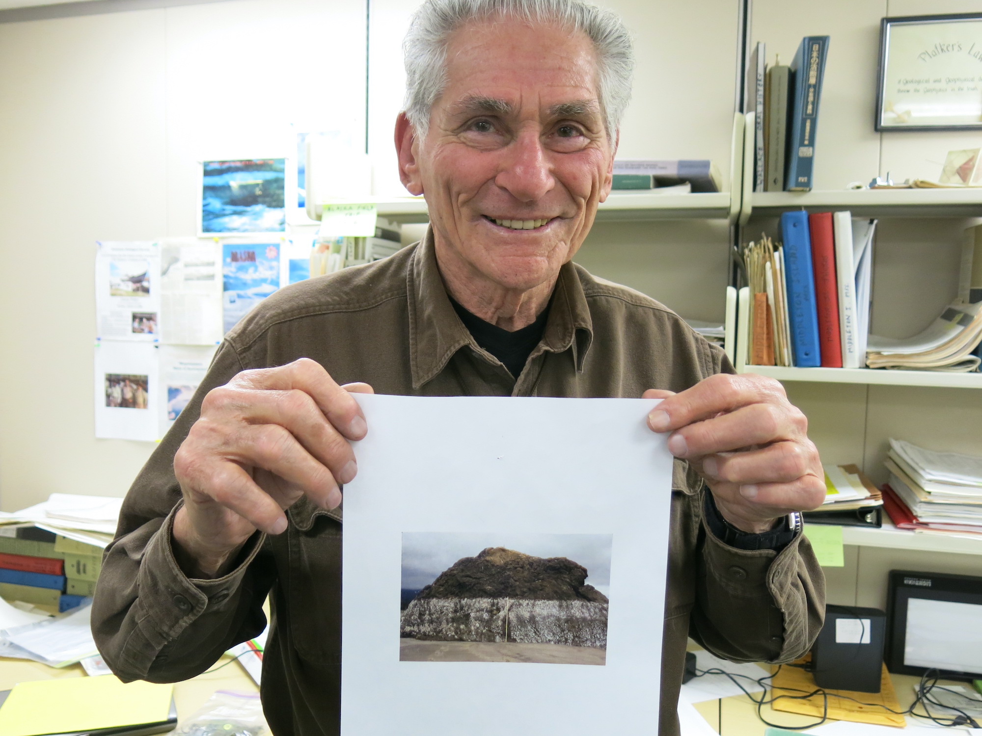 A man holds a photograph of a large seaside outcrop, the bottom half of which has turned white from desiccated barnacles and other shoreline creatures. 