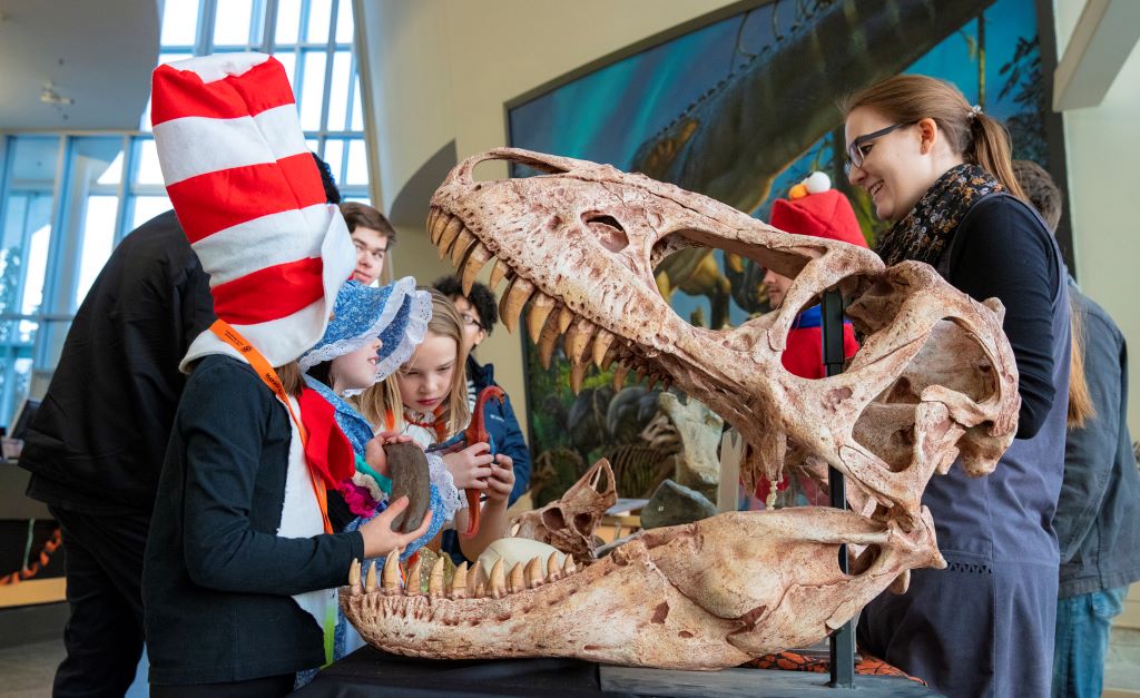 Children in Halloween costumes handle fossils and look at a large dinosaur skull in a museum, with a smiling adult.