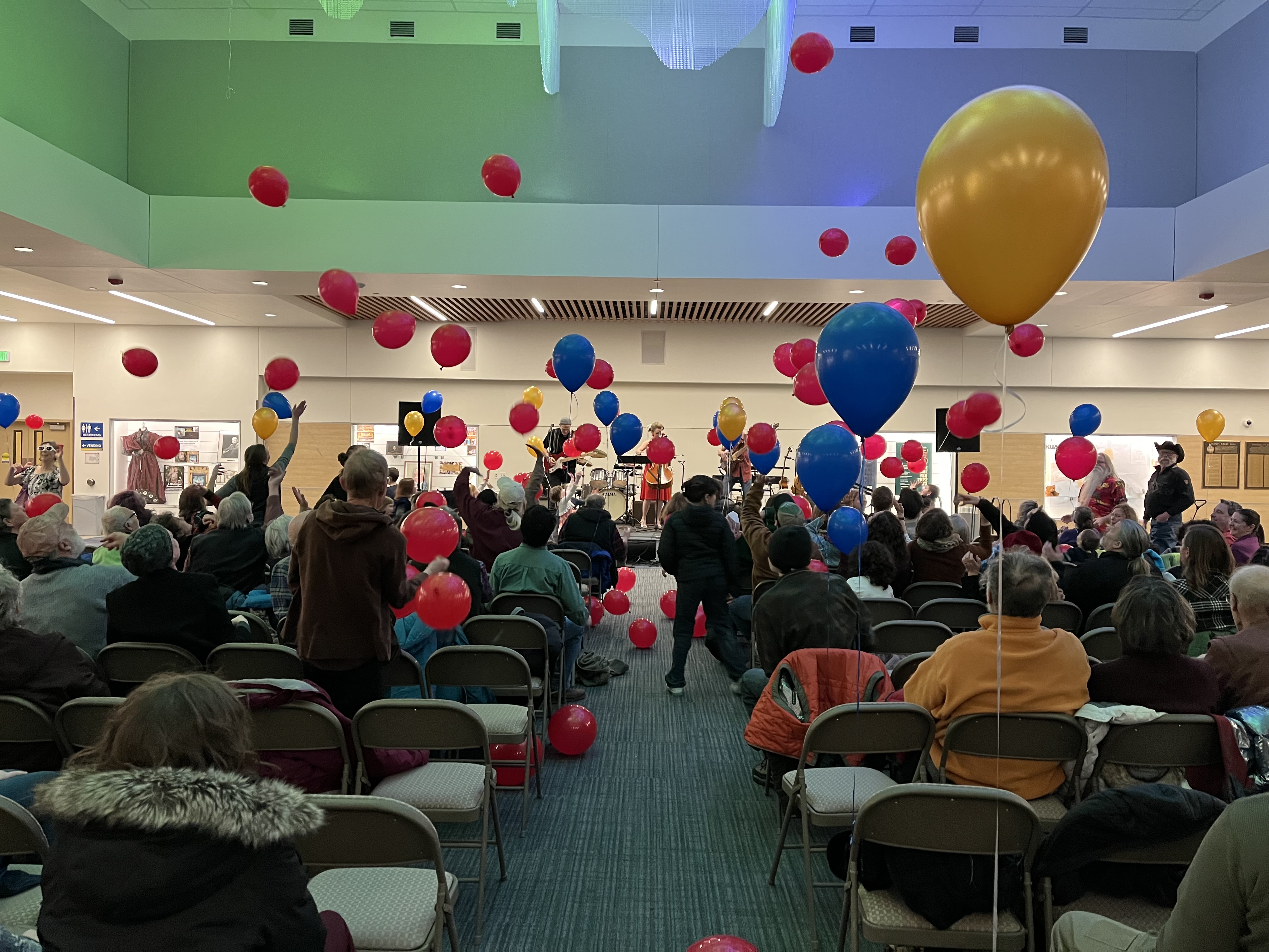 photo shot from the back of an audience towards a stage, with blue, gold and red helium balloons