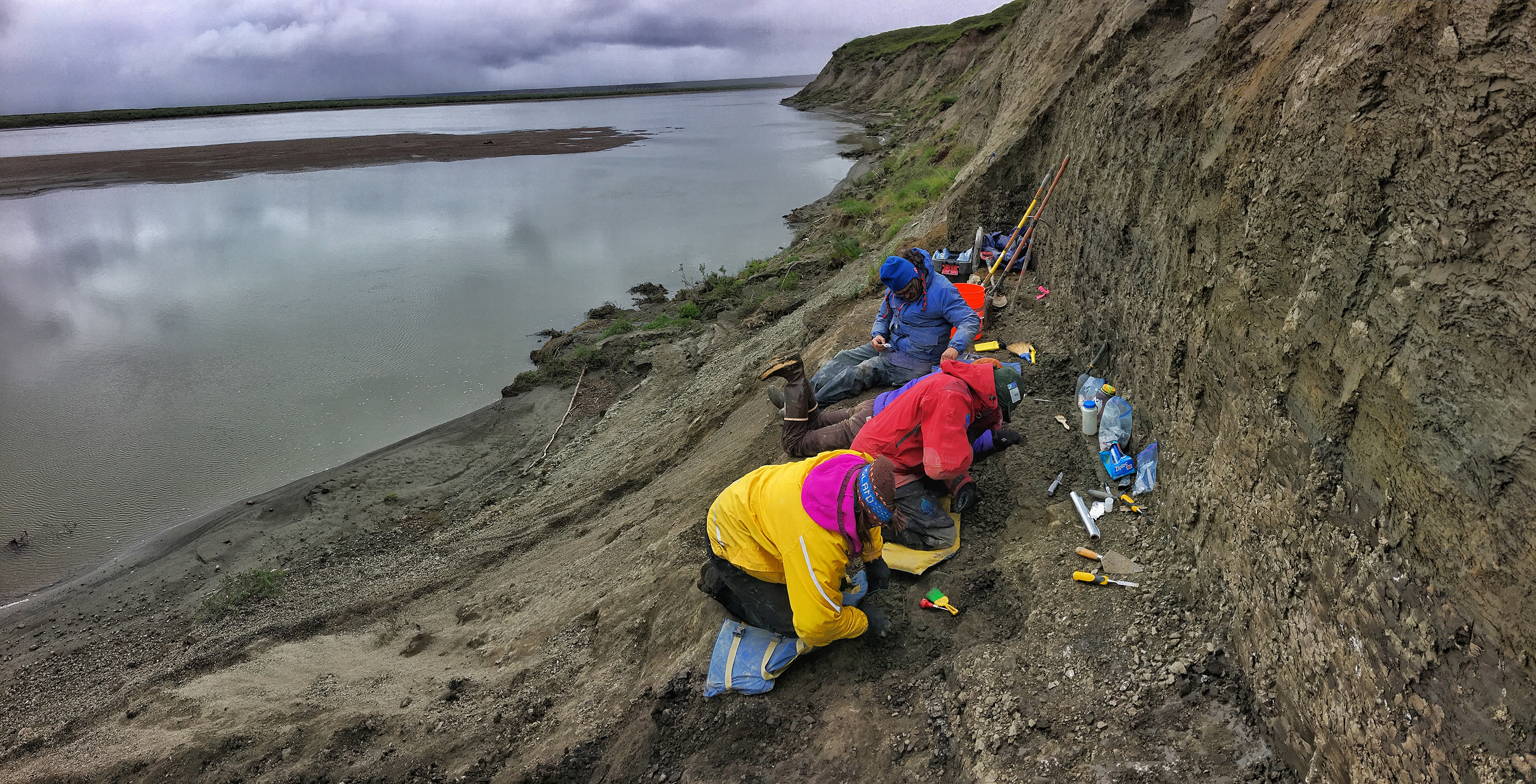 Three people in outdoor clothing dig in dirt on the side of a cliff with a large river in the background. 