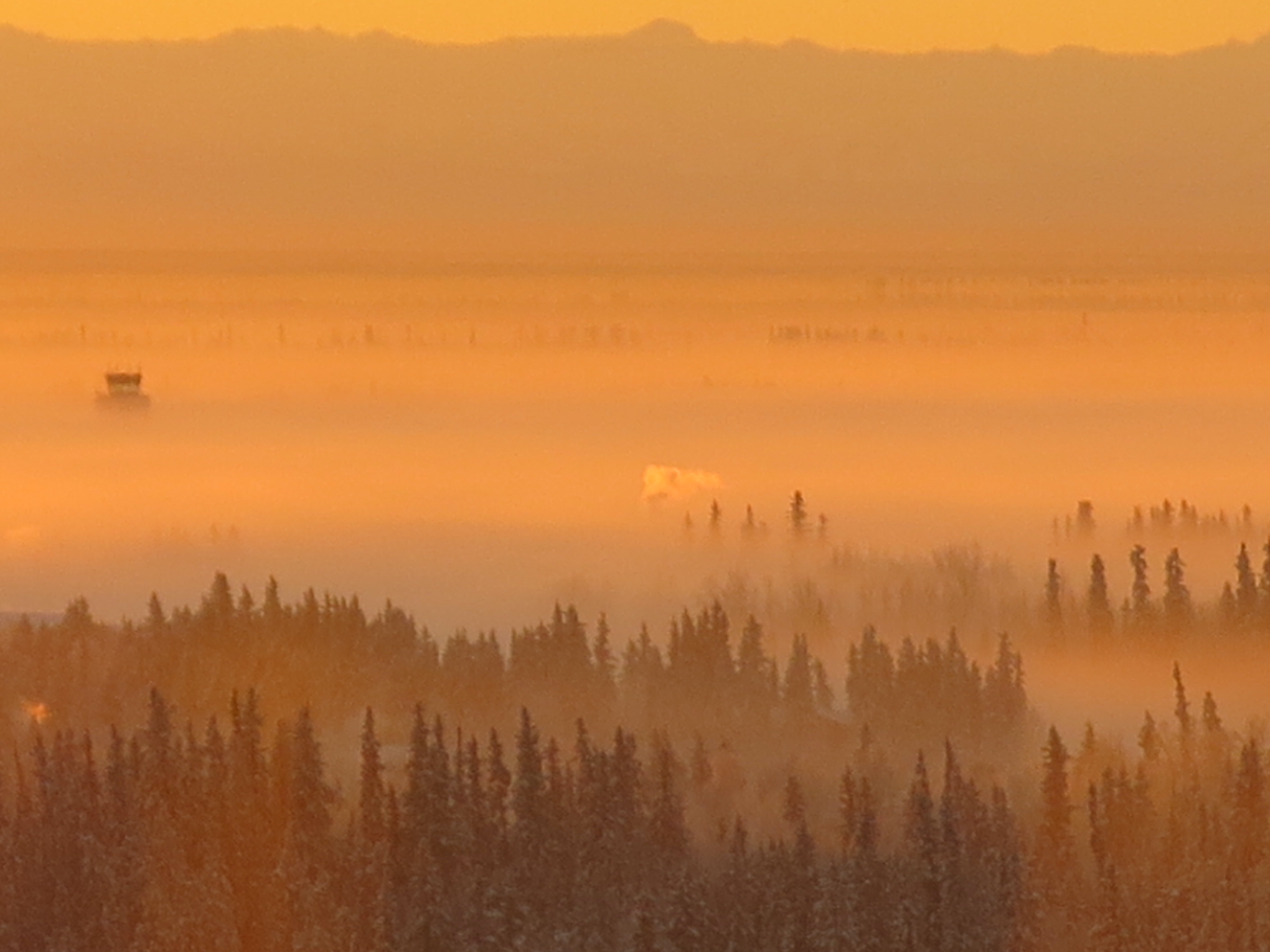 Orange mid-winter light bathes a flatland covered in a low-lying fog. The tops of spruce trees and of an airport control tower poke up through the fog. Mountains form a distant rugged horizon. 