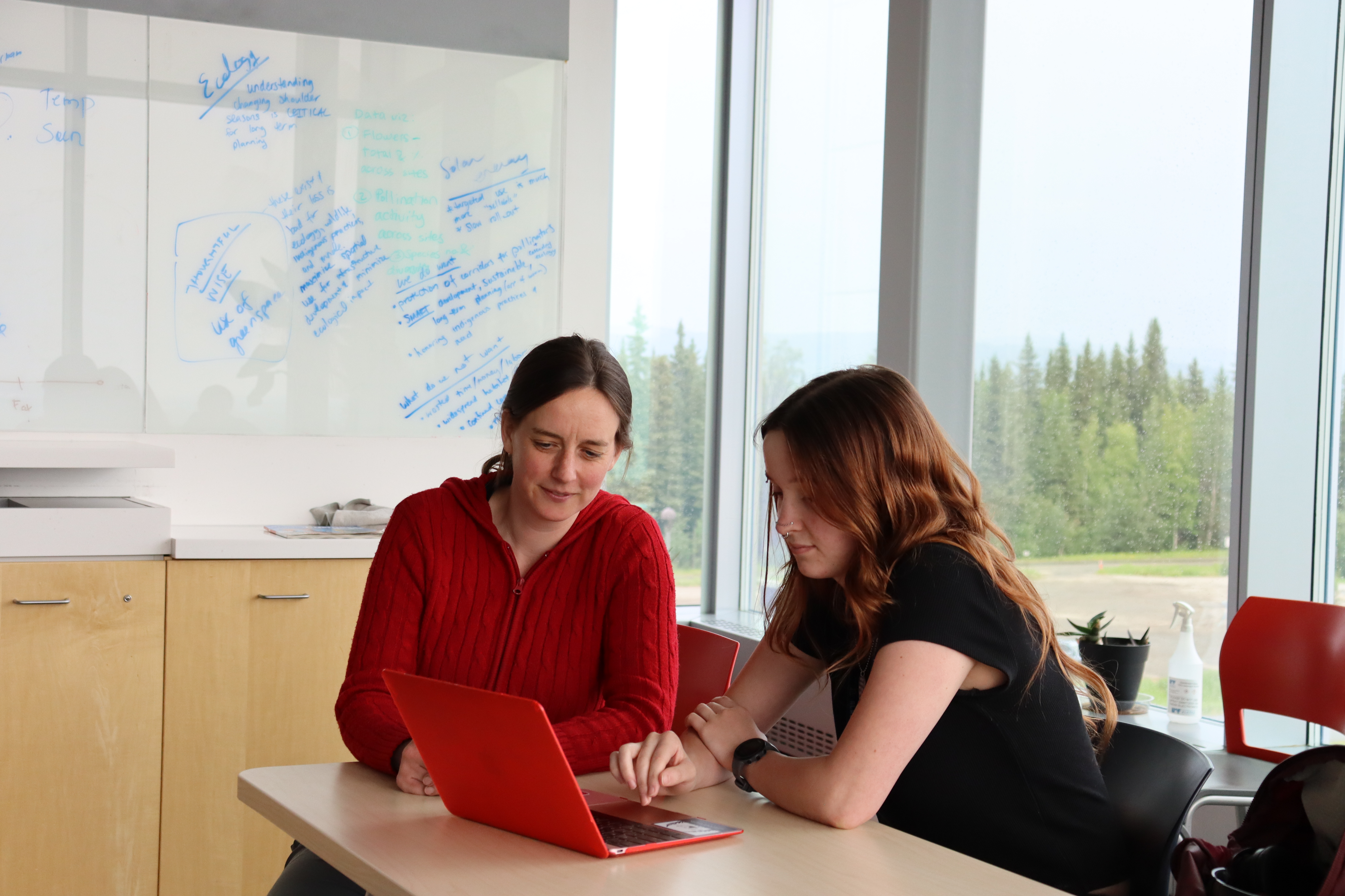 In a classroom with a whiteboard full of notes, an instructor and student look at a laptop together.