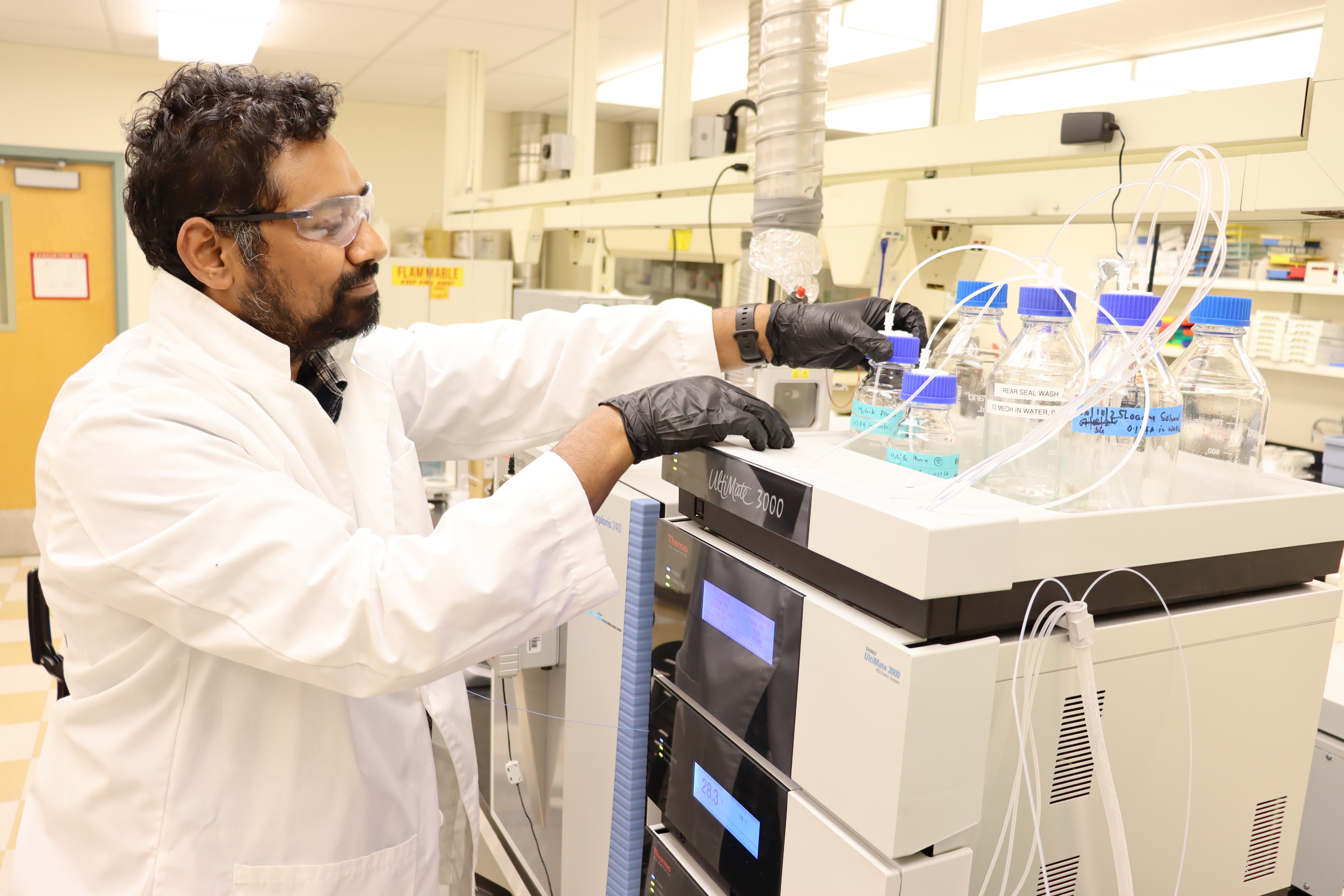 A man in a white lab, safety glasses and rubber gloves places a glass container in a tray atop a large piece of lab equipment.