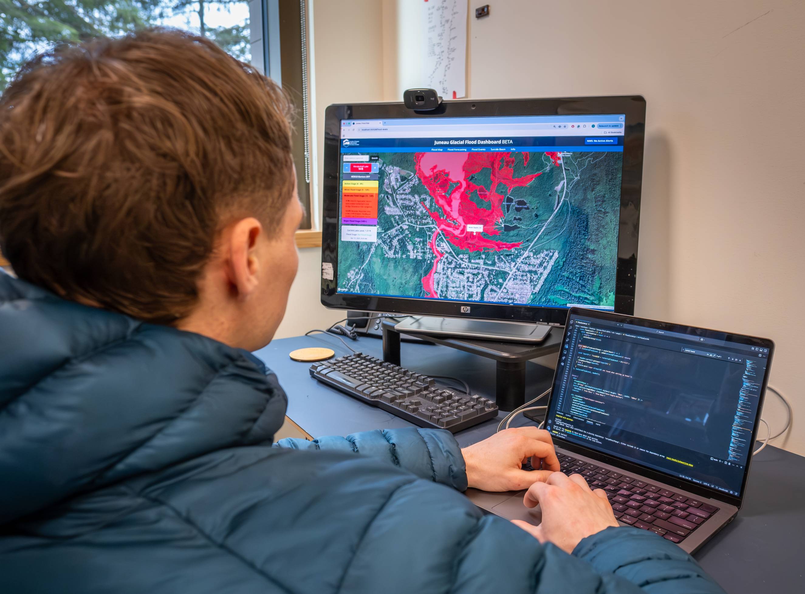 Photographed from behind, a person works on a small laptop beside a large screen showing a map of Juneau overlaid with large red area indicating a flooding scenario.
