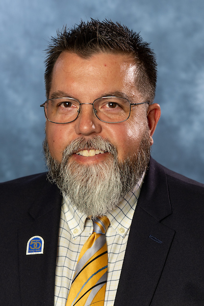 A man wears a black suit jacket, a patterned shirt, and tie for a portrait.