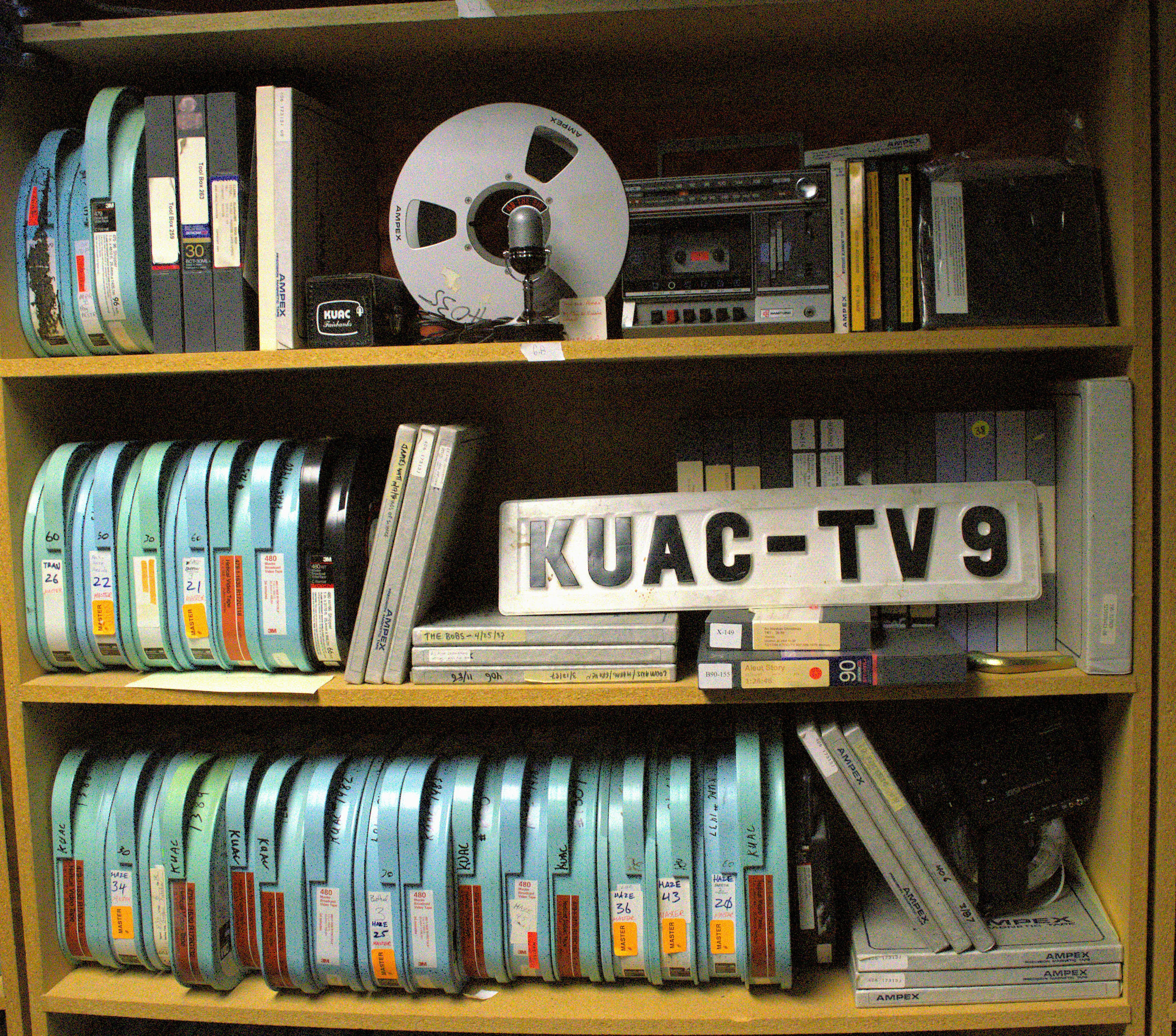 Tape reels, video film canisters, a tape deck and other materials sit on three shelves along with an old sign for 