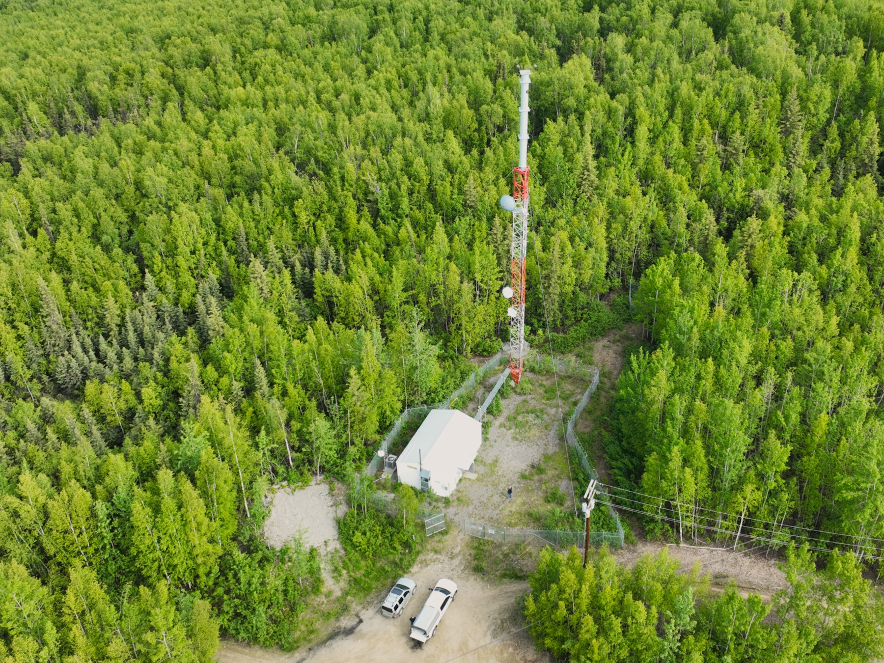 Overhead view of a transmitter tower and building in a clearing surrounded by trees in summer foliage