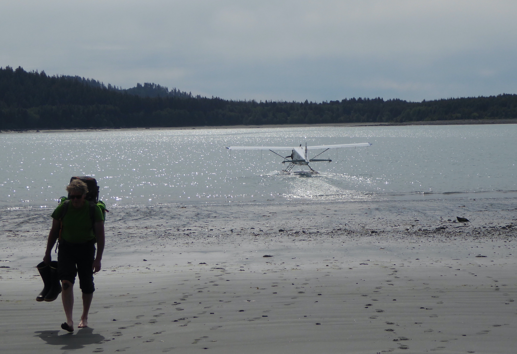 A barefoot man walks up a beach while a floatplane leaves the shoreline behind him.