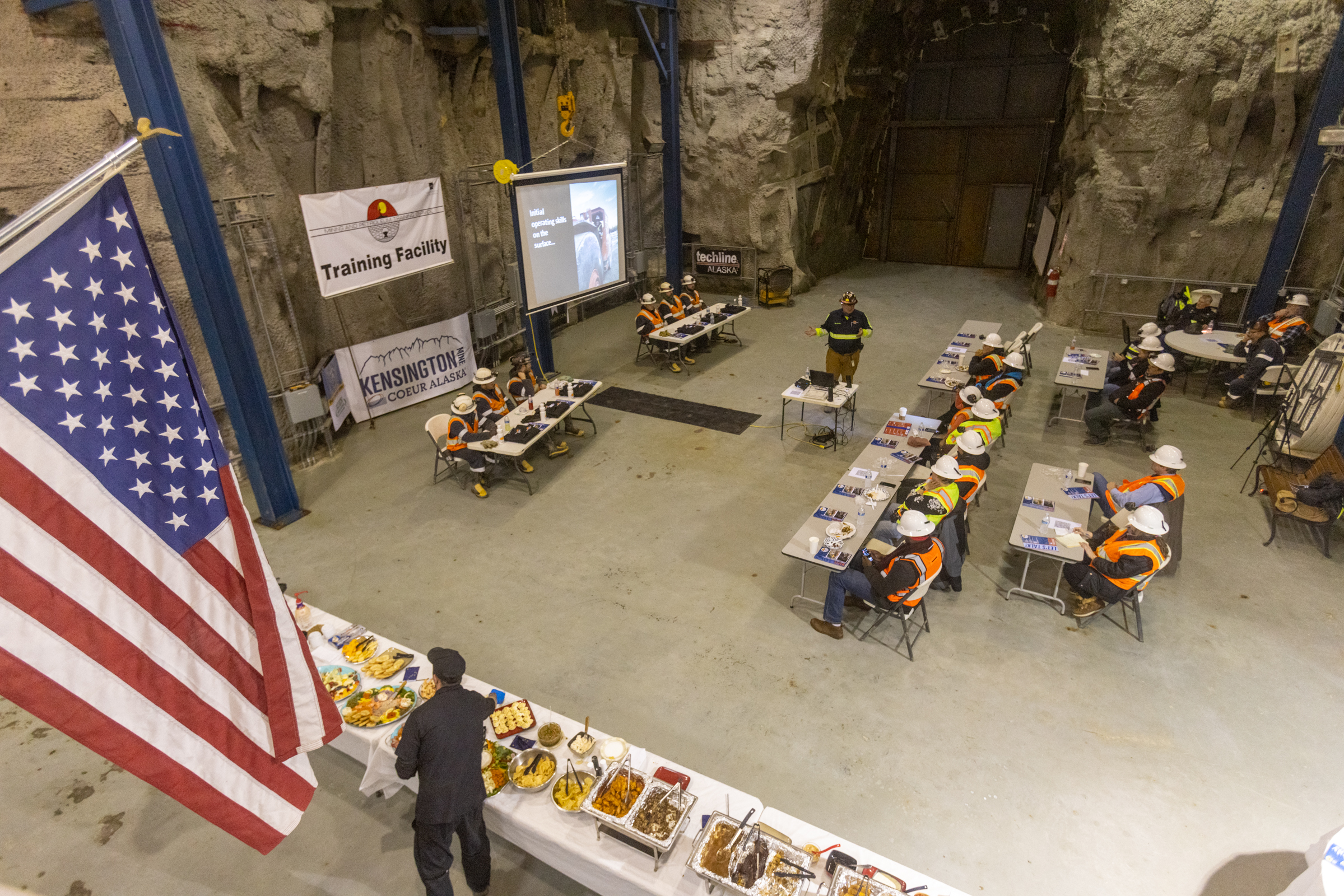people wearing hard hats and safety vests seated in chairs looking at a presentation, as viewed from above.