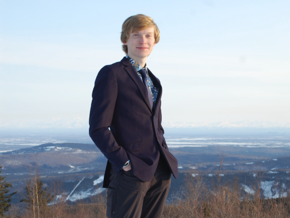 A man in a suit stands on a high hill with a view down to a valley.