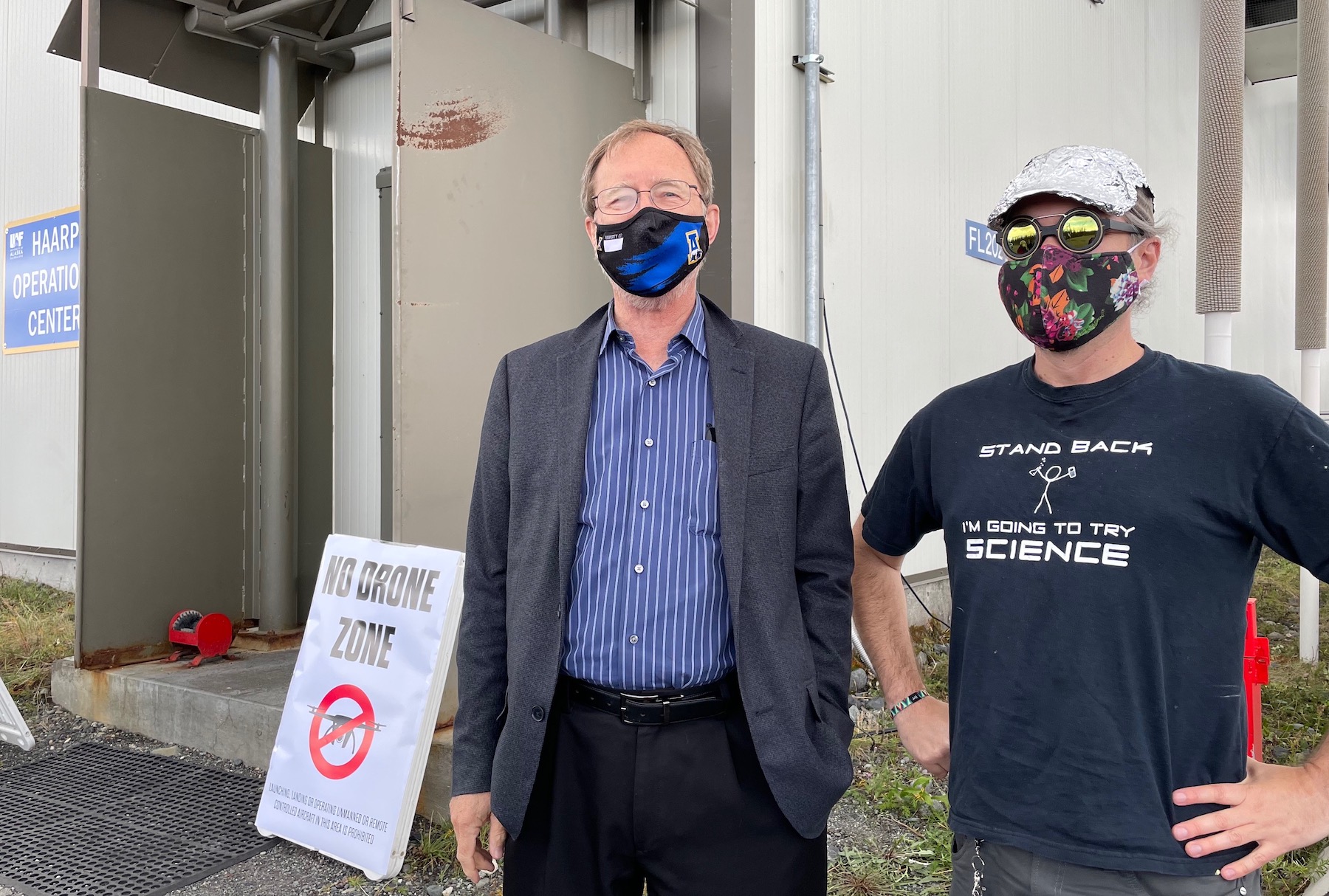 Two men in COVID-19 facemasks stand in front of a white and gray metal building. The man on the left wears a dress shirt and slacks and a sport coat.  The man on the right wears a ball cap covered with tinfoil, reflective round sunglasses and a T-shirt that says "Stand back. I'm going to try science."
