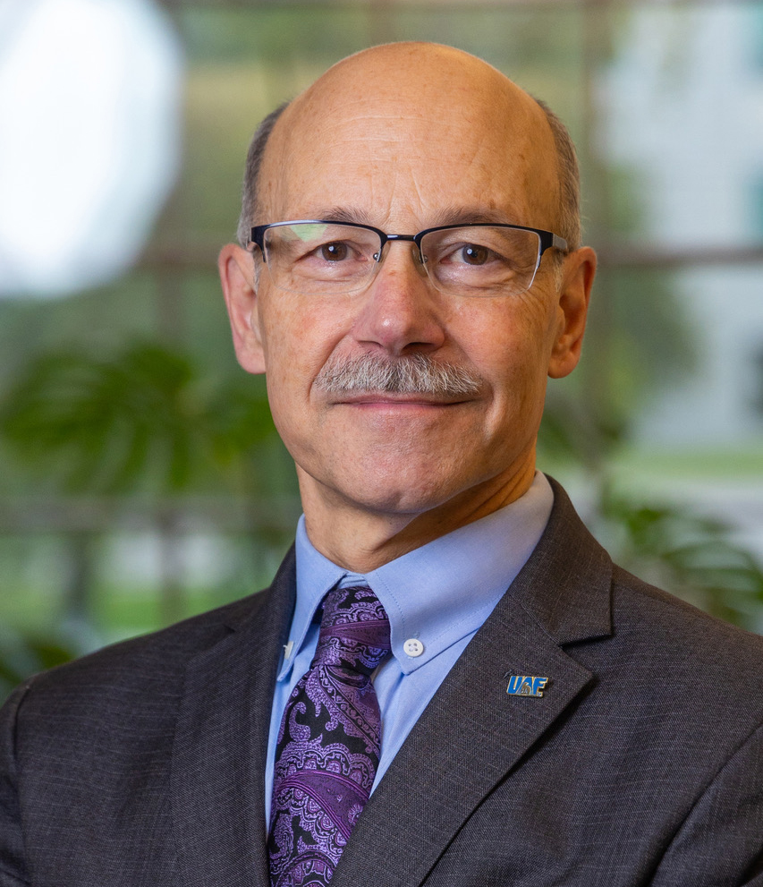 A man wearing glasses, a dark gray business jacket, a light blue button-up shirt, a purple and black swirl pattern tie, and a UAF pin on his lapel.