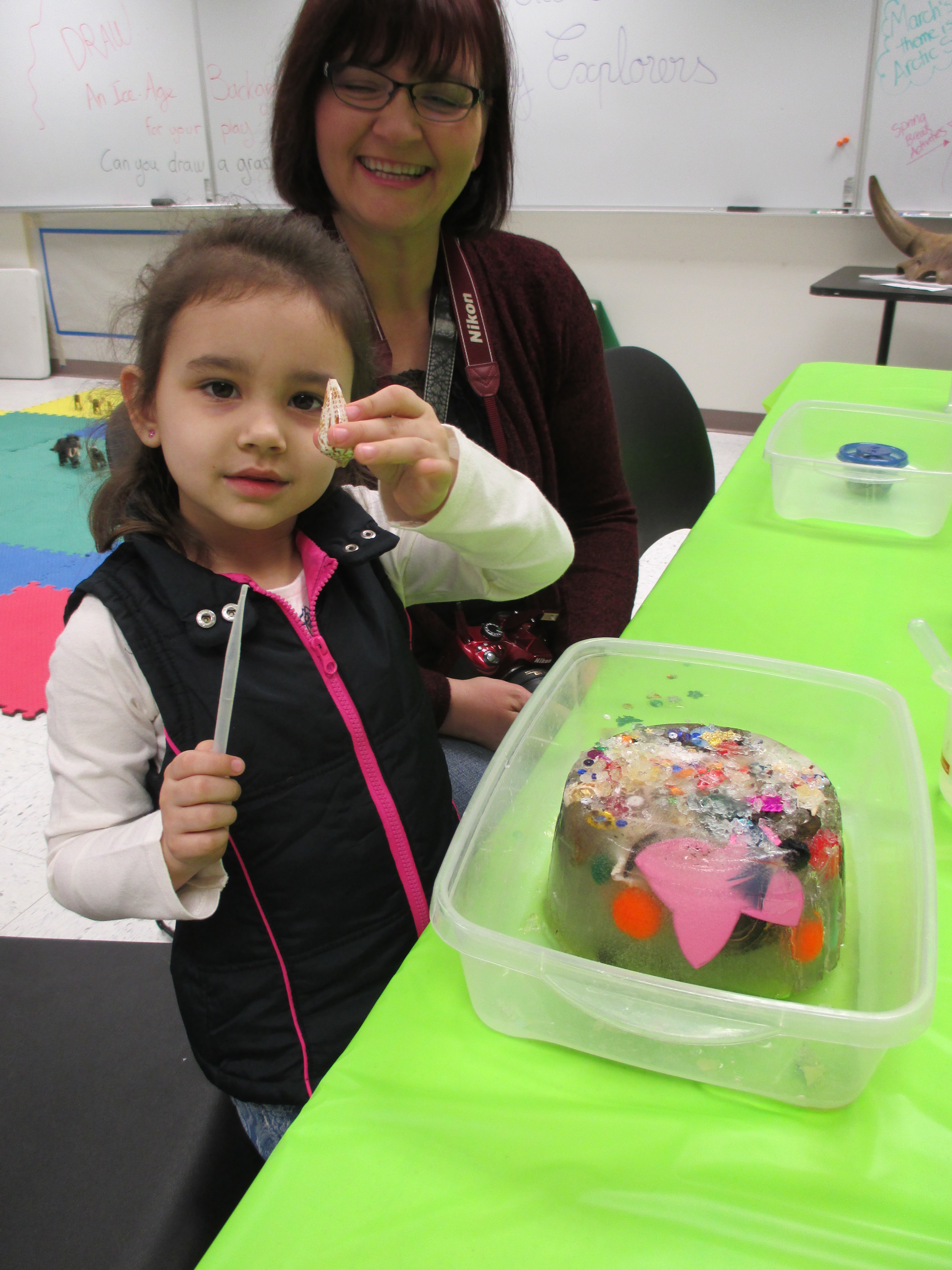 A young girl holds up a seashell. An adult sits behind her, smiling. On the table in front of her is a plastic bin containing a block of ice with beads, balls and many other items frozen inside it.