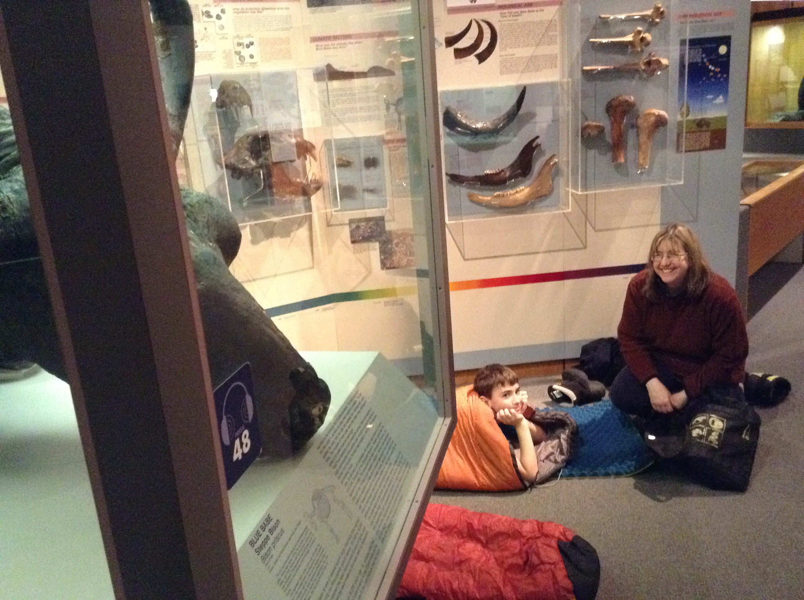 a woman sitting on the floor of a museum beside a child in a sleeping bag, surrounded by display cases