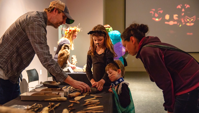 Two children in Halloween costumes and two adults look together at museum artifacts on a table. Images of Jack-o'-lanterns are projected on the wall behind them.